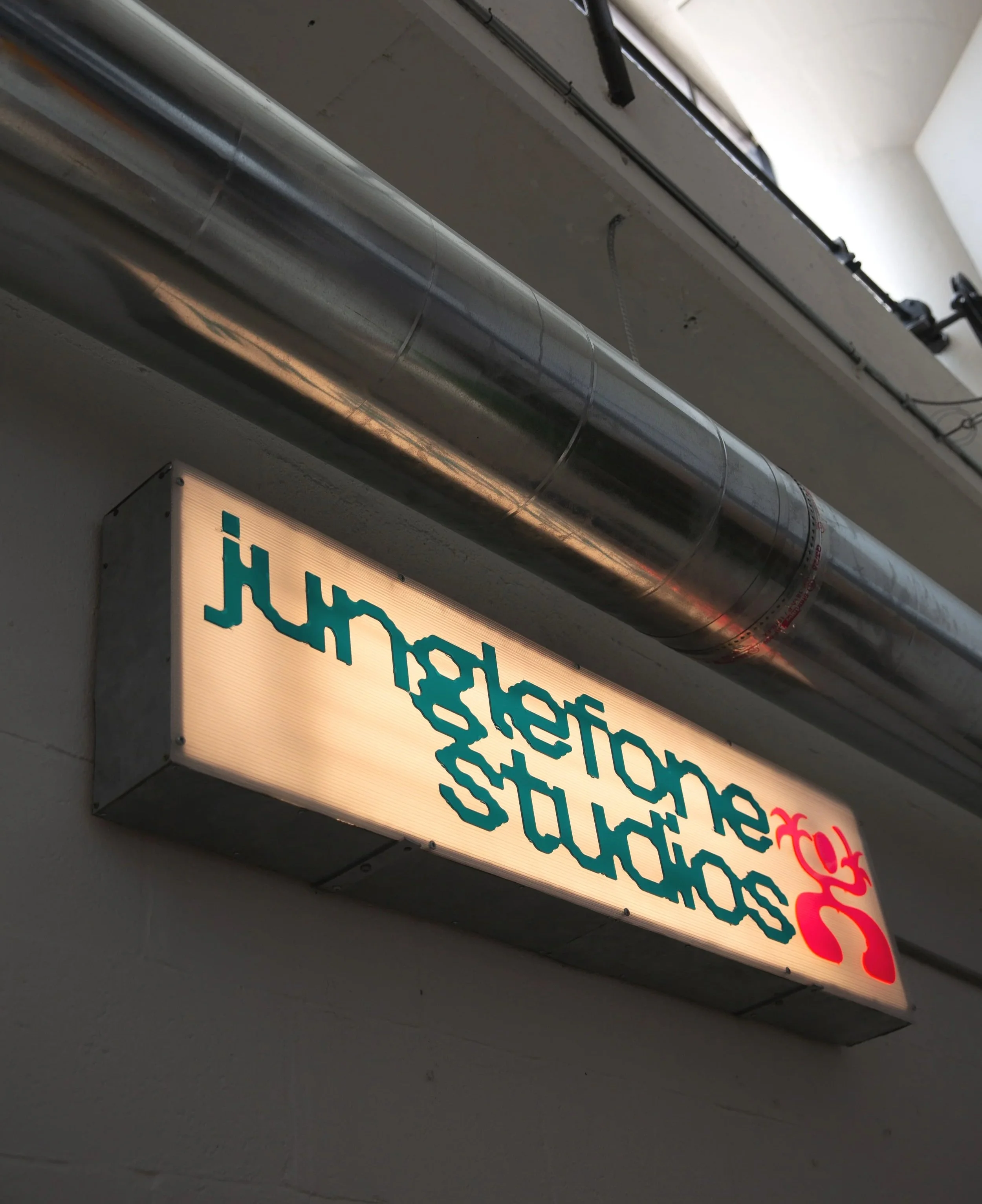 Illuminated sign with partially visible text in a warehouse or industrial setting, showing pipes and electrical conduits on the ceiling.