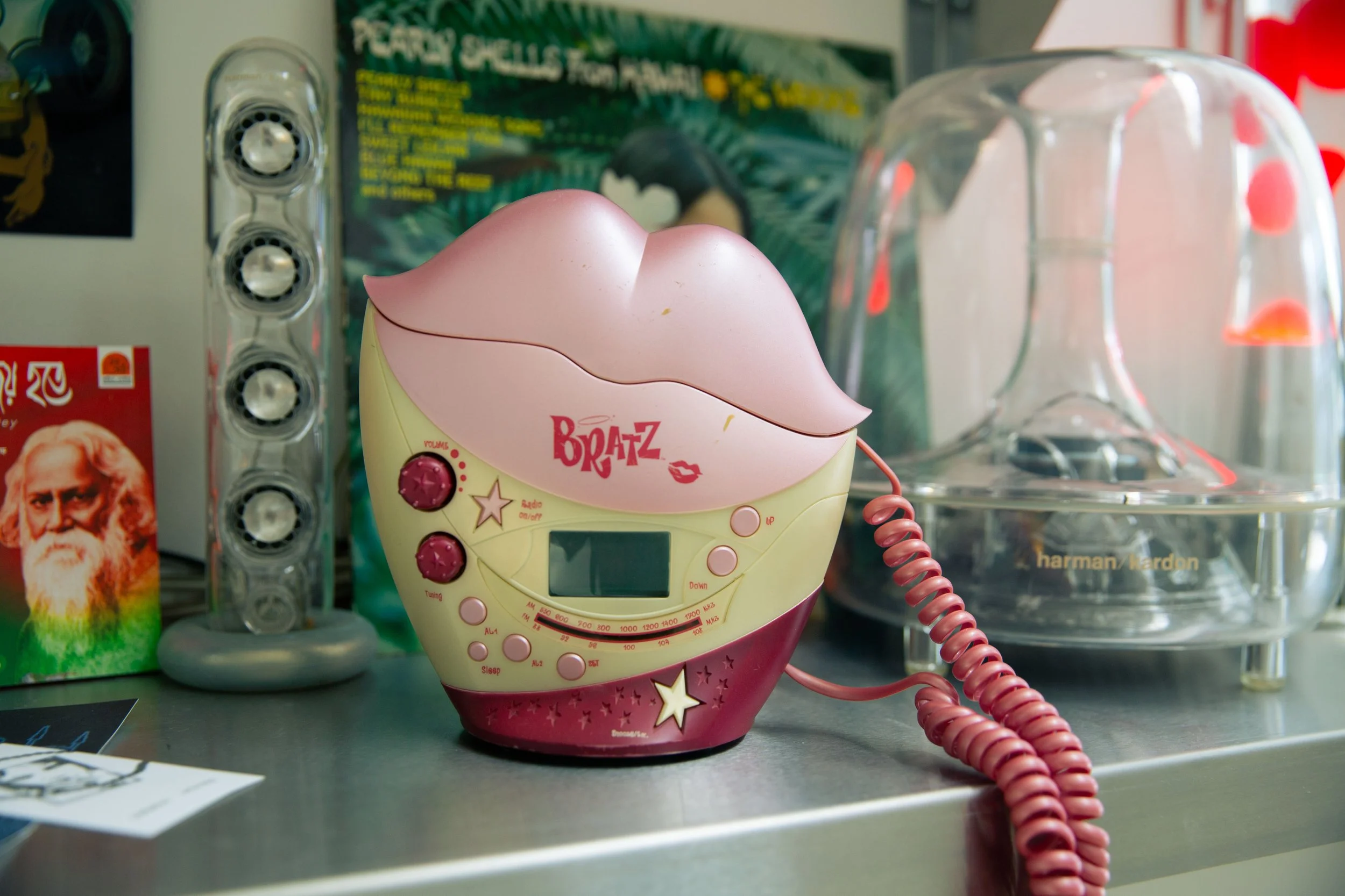 A pink and yellow toy karaoke machine with a comic lips design on the front, sitting on a green surface, with a pink coiled cord connected to the microphone. Behind it, there are various objects including a clear plastic container with orange and pin
