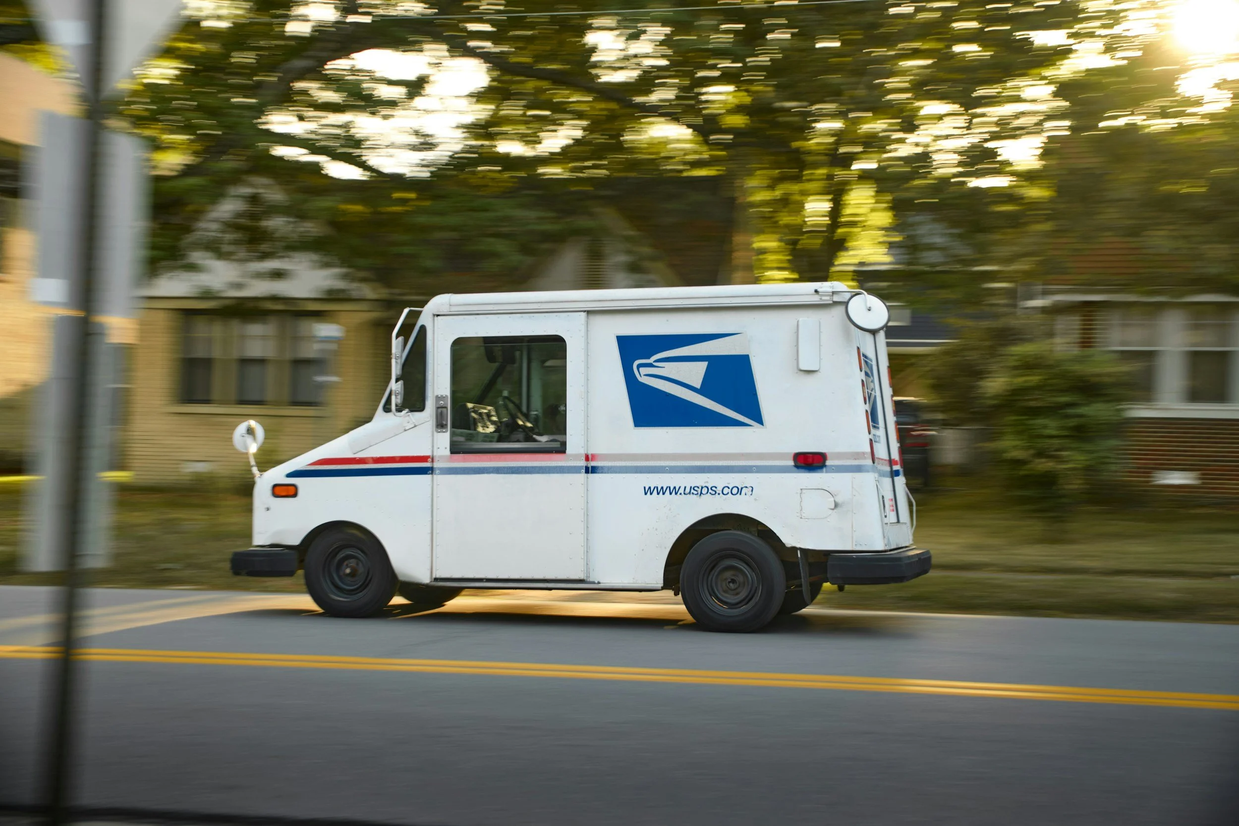 A USPS postal delivery truck is driving on a suburban street with houses and trees in the background. The photo appears to be taken in motion, creating a blurred effect.