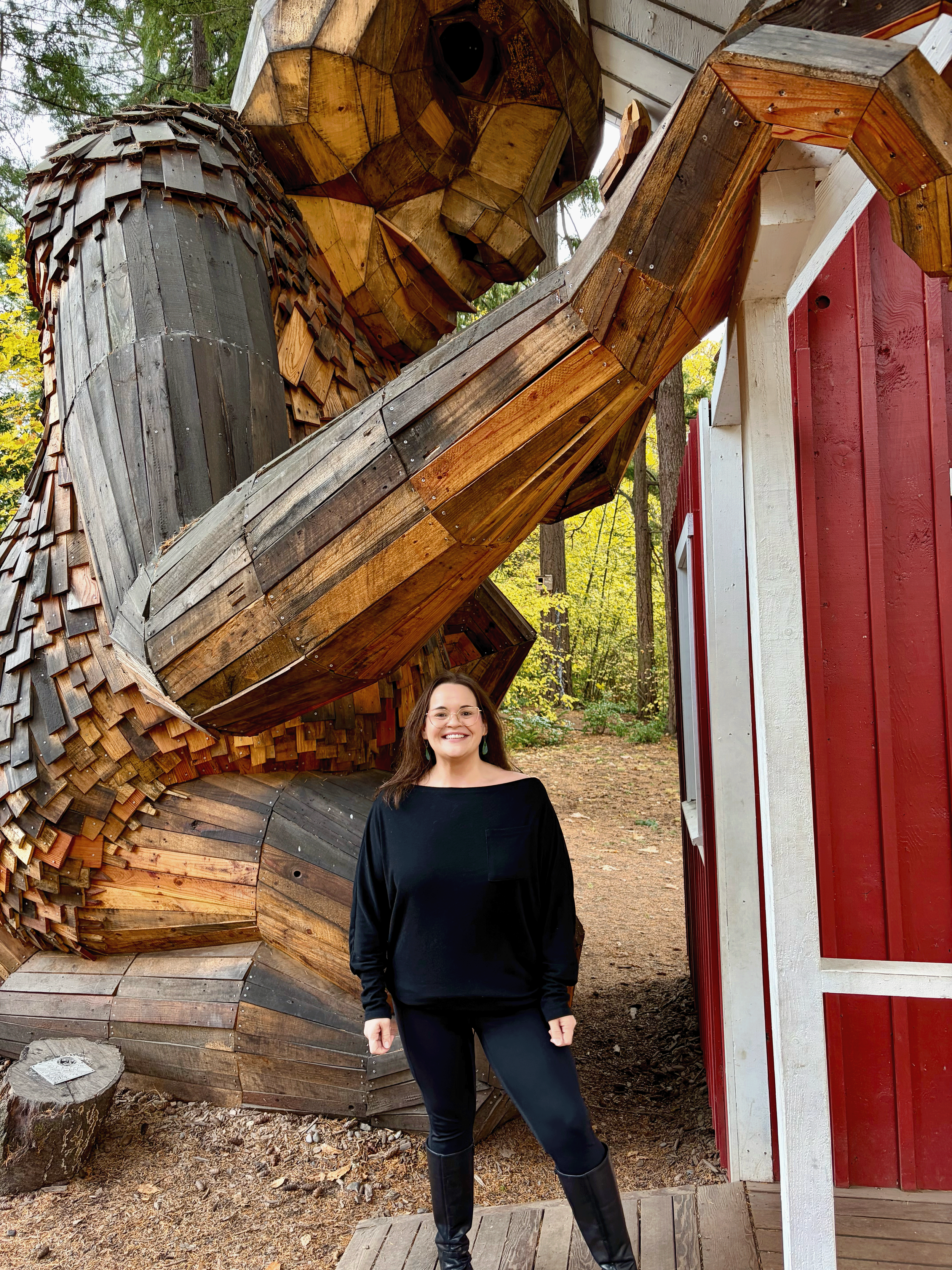 Woman smiling, standing outdoors near a red and white wooden building with a large sculpture made of wood in the background.
