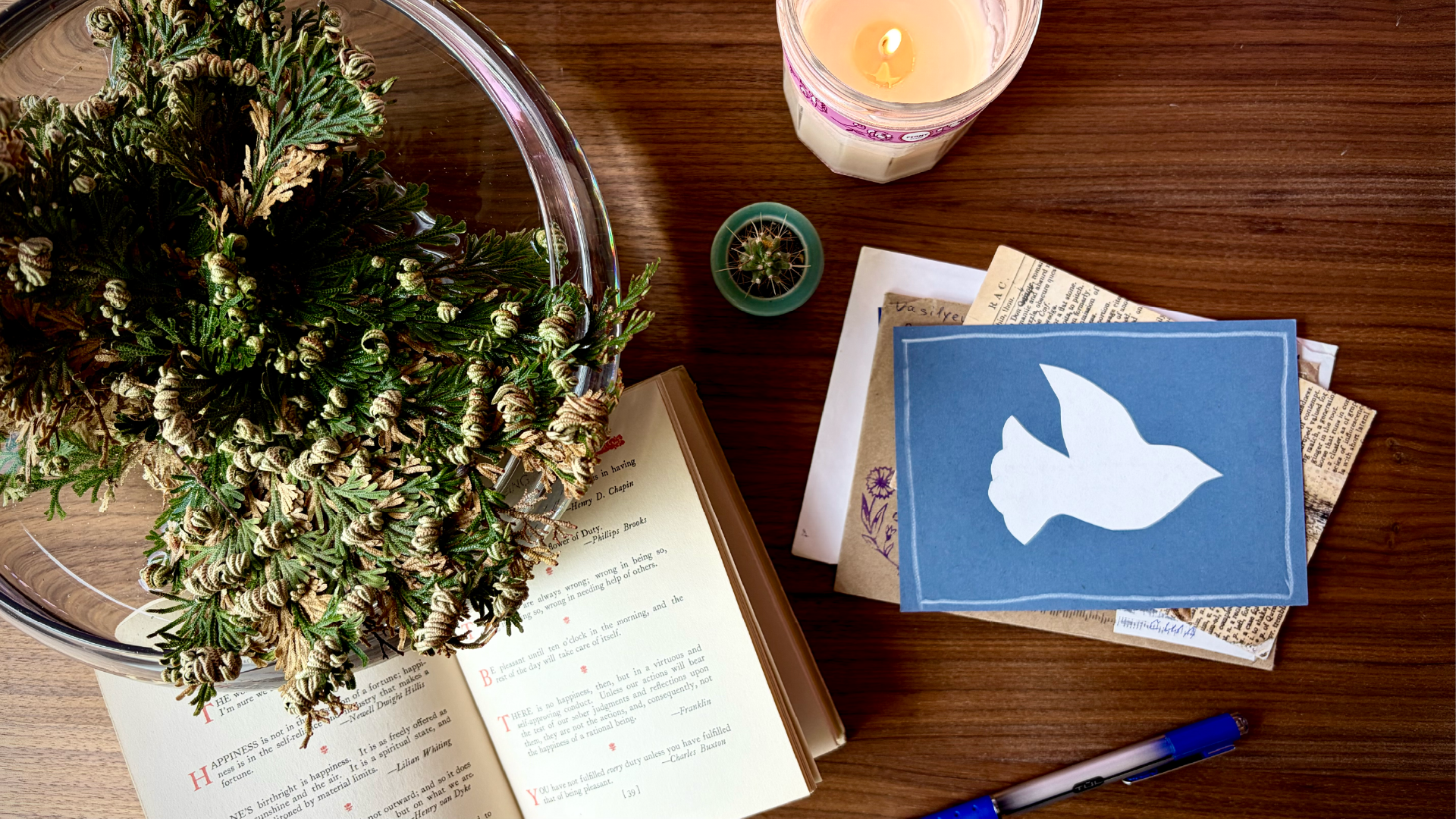 A wooden table with a large potted plant, a candle, a book opened to a page, a blue card with a white dove cutout, several newspapers, and a blue and black pen.