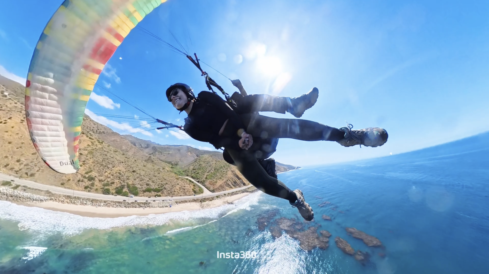 Persona practicando parapente sobre la playa y el mar en un día soleado, con montañas al fondo.