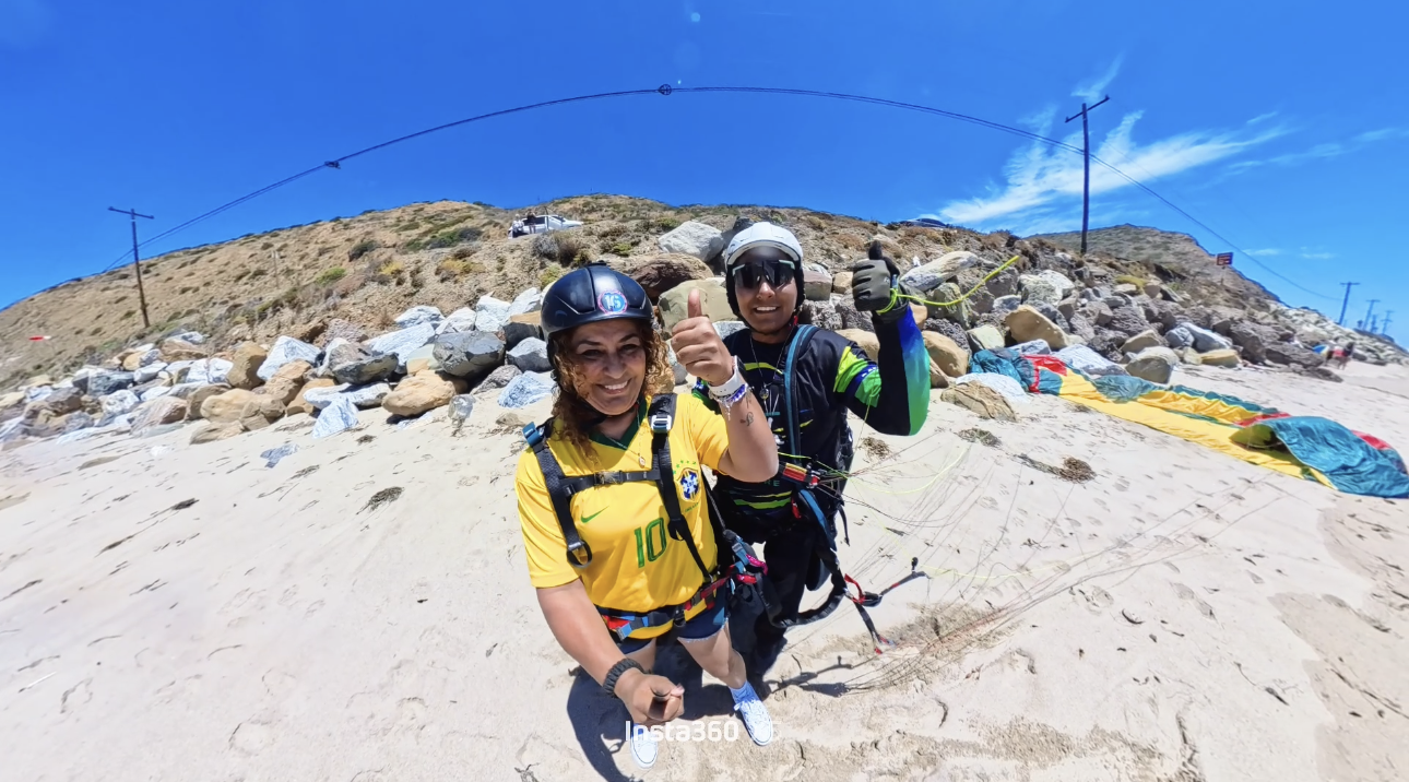 Dos personas en equipo de parapente en la playa, sonriendo y haciendo un gesto de aprobación, con cielo azul y rocas en el fondo.
