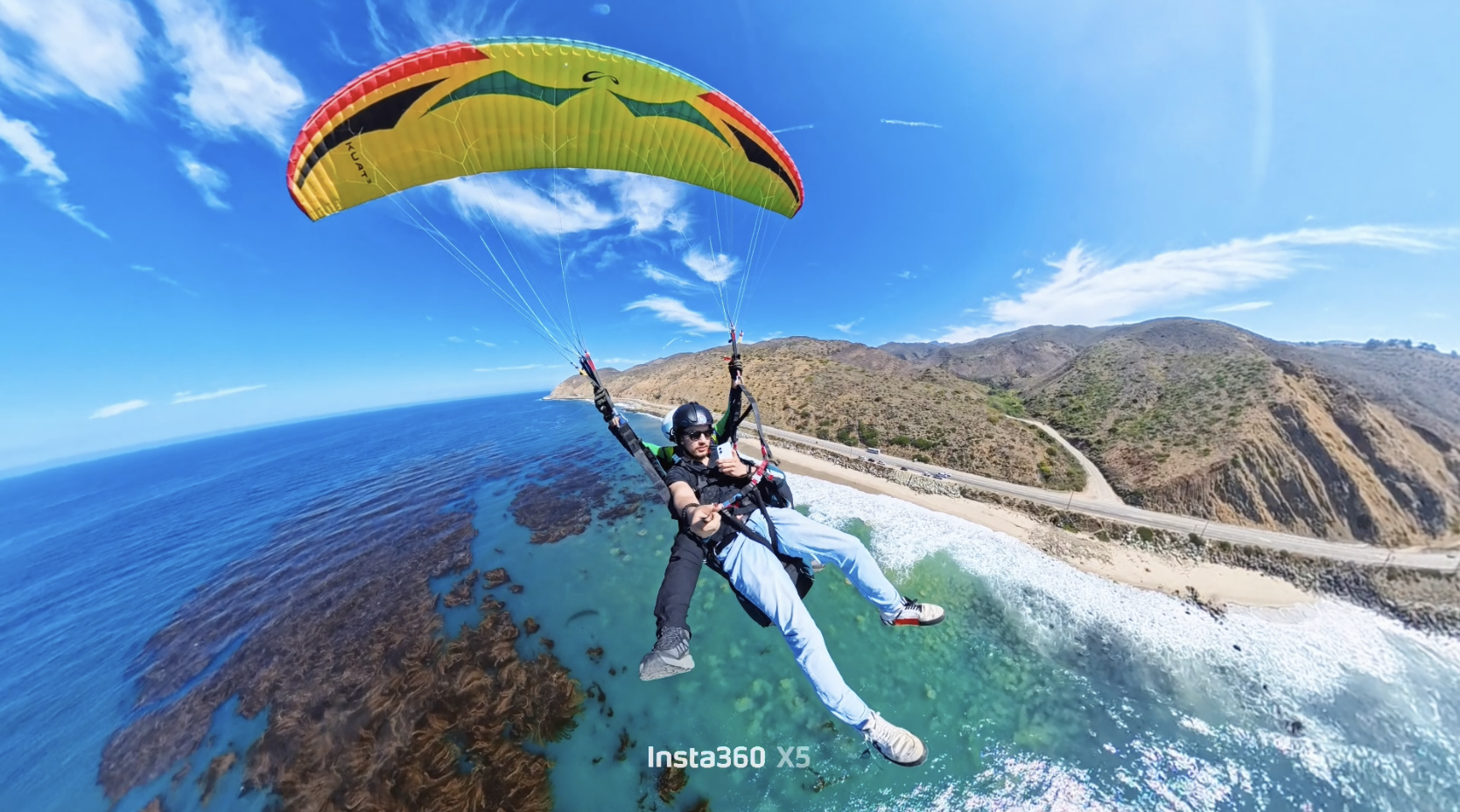 Persona practicando parapente sobre la playa con montañas y océano al fondo en un día soleado.