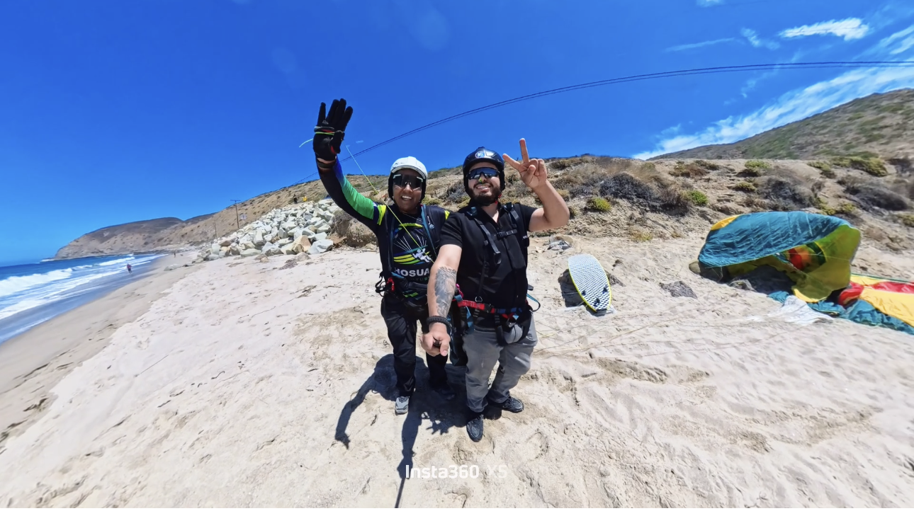 Dos personas con equipo de paracaidismo en la playa, sonriendo y haciendo gestos de paz, con paracaídas desplegado sobre la arena y el mar en el fondo.