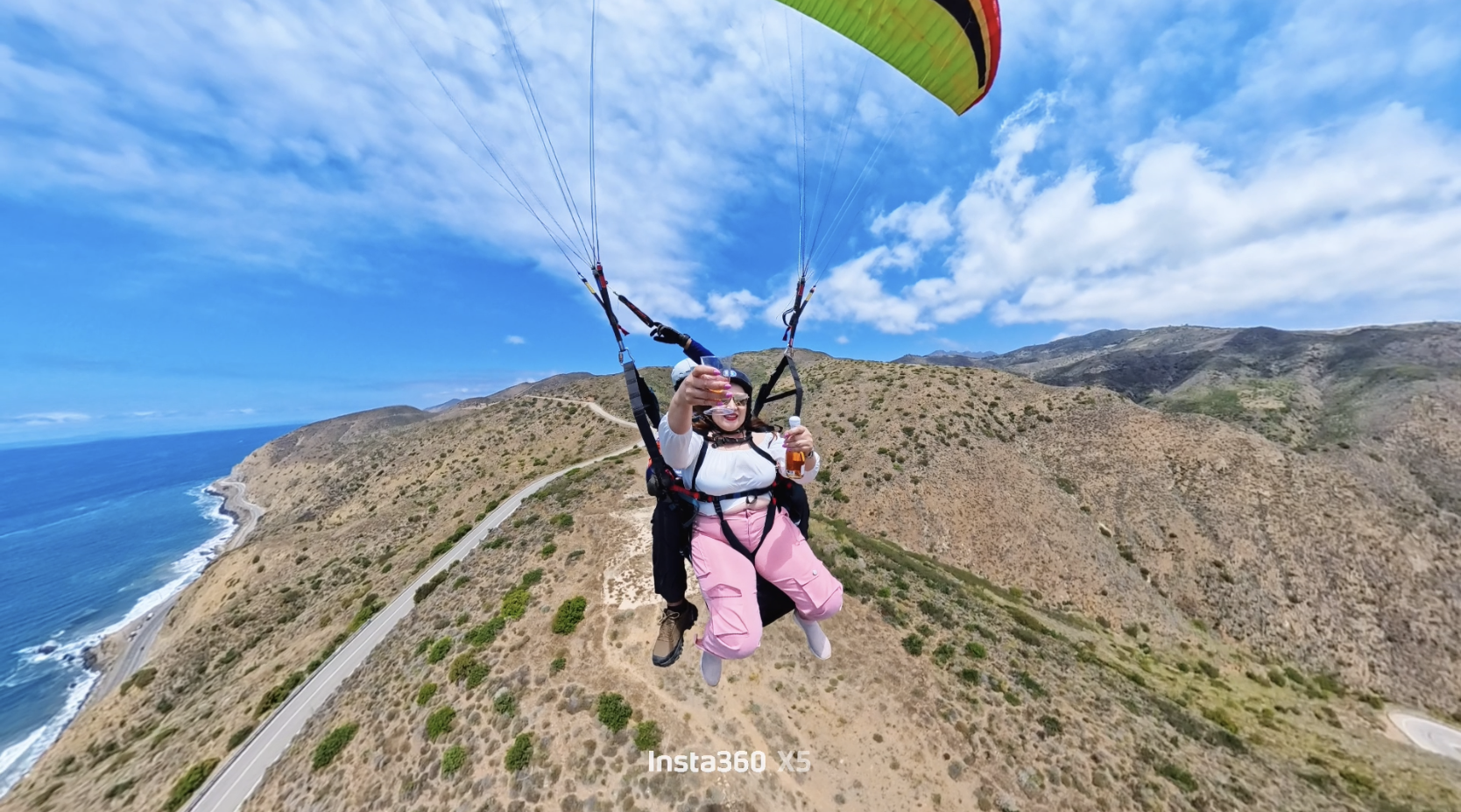 Persona haciendo parapente sobre la costa con montañas en el fondo, tiende una copa y una botella en la mano.