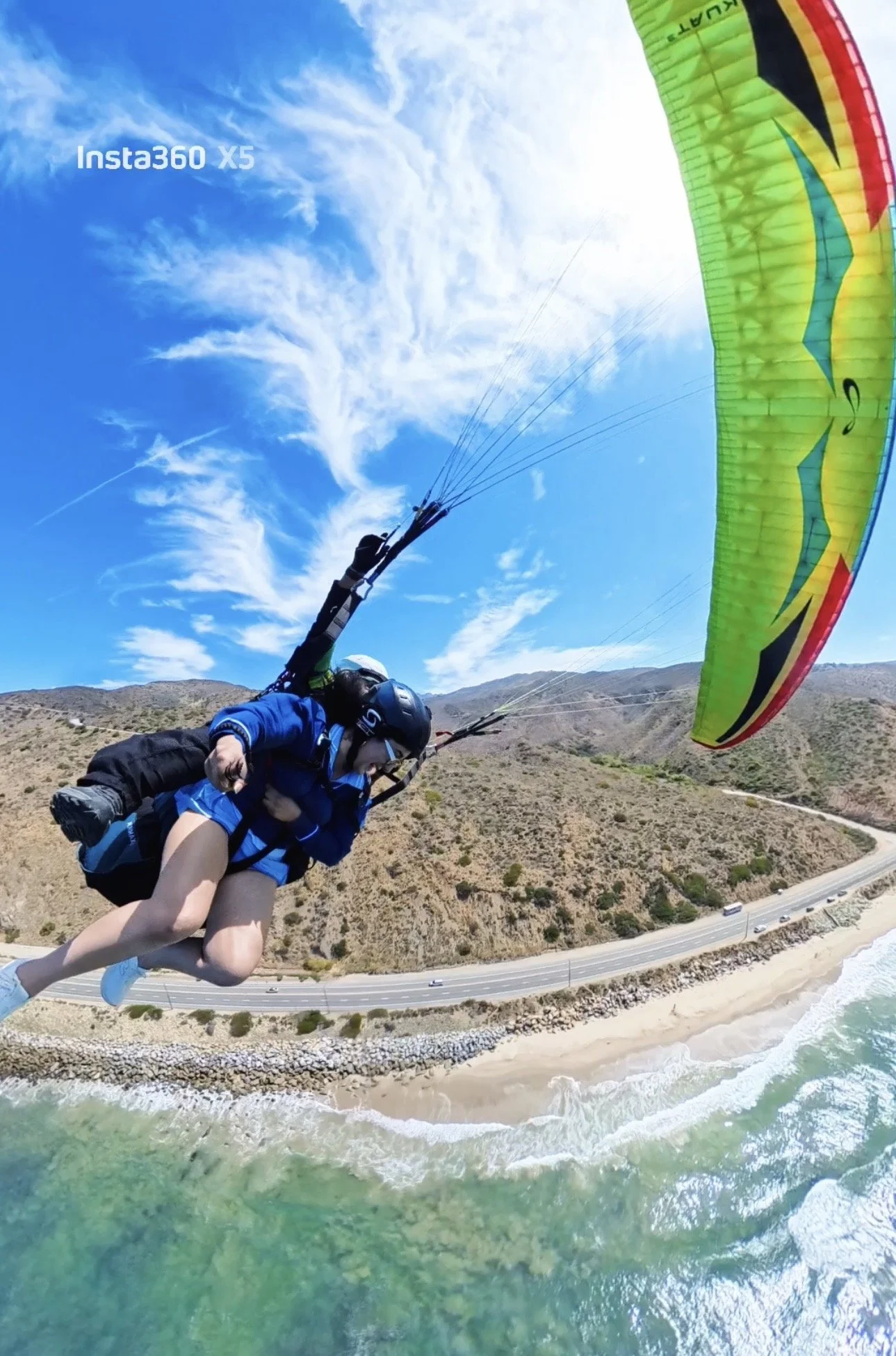 Persona practicando parapente sobre una playa con montañas y carretera, en un día soleado con cielo despejado.