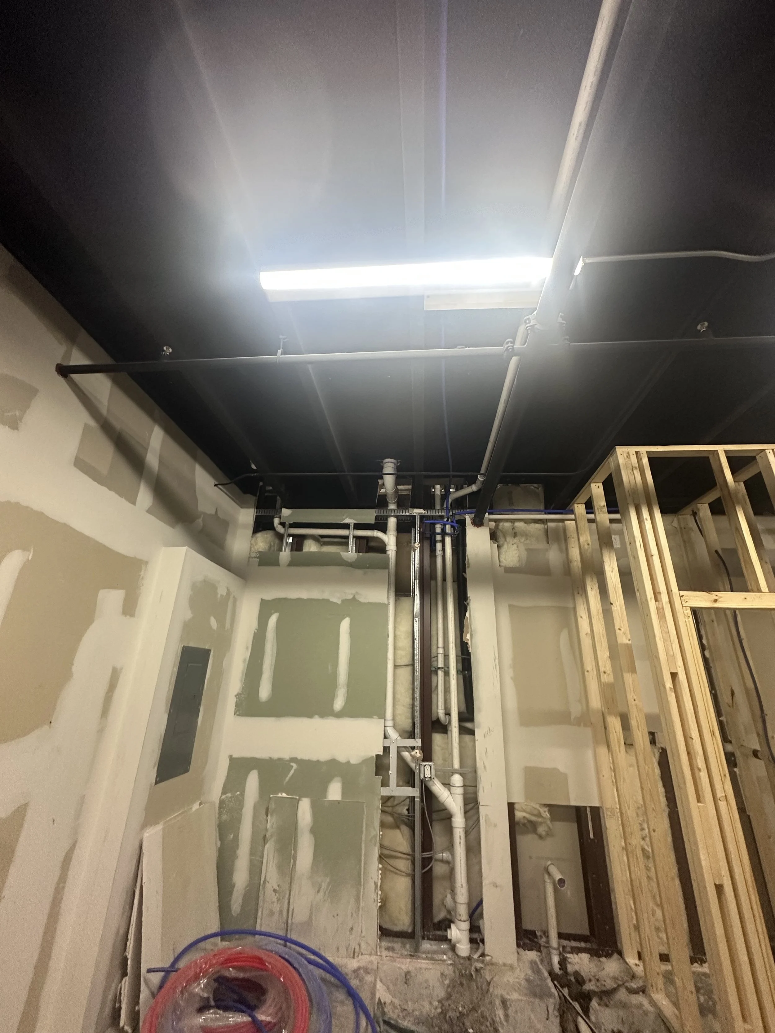 Construction area with open wall, exposed pipes, wooden framing, and construction materials.