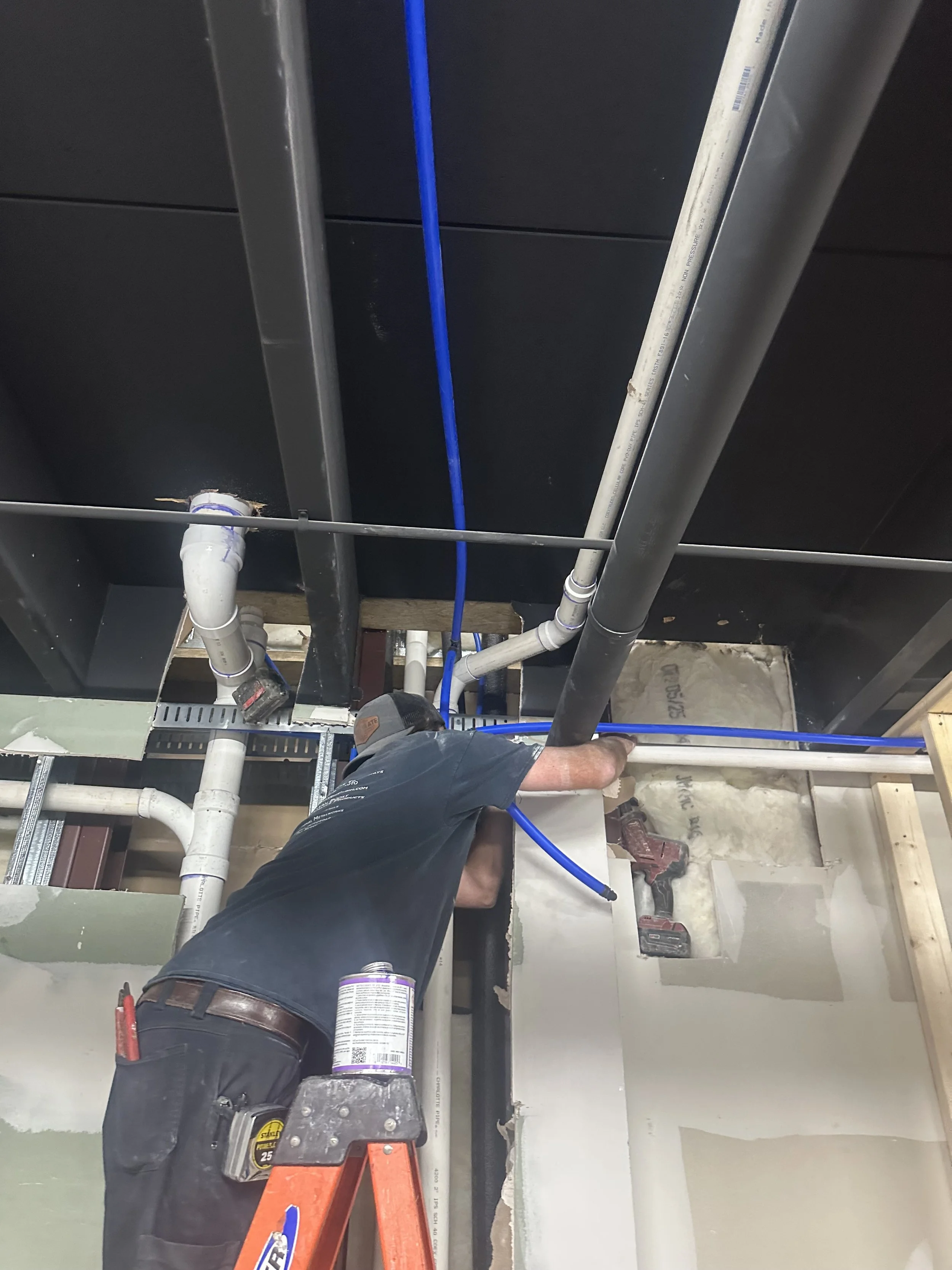 A worker installing or repairing plumbing pipes in a ceiling space. The worker is standing on a small ladder and working with various pipes and tools.