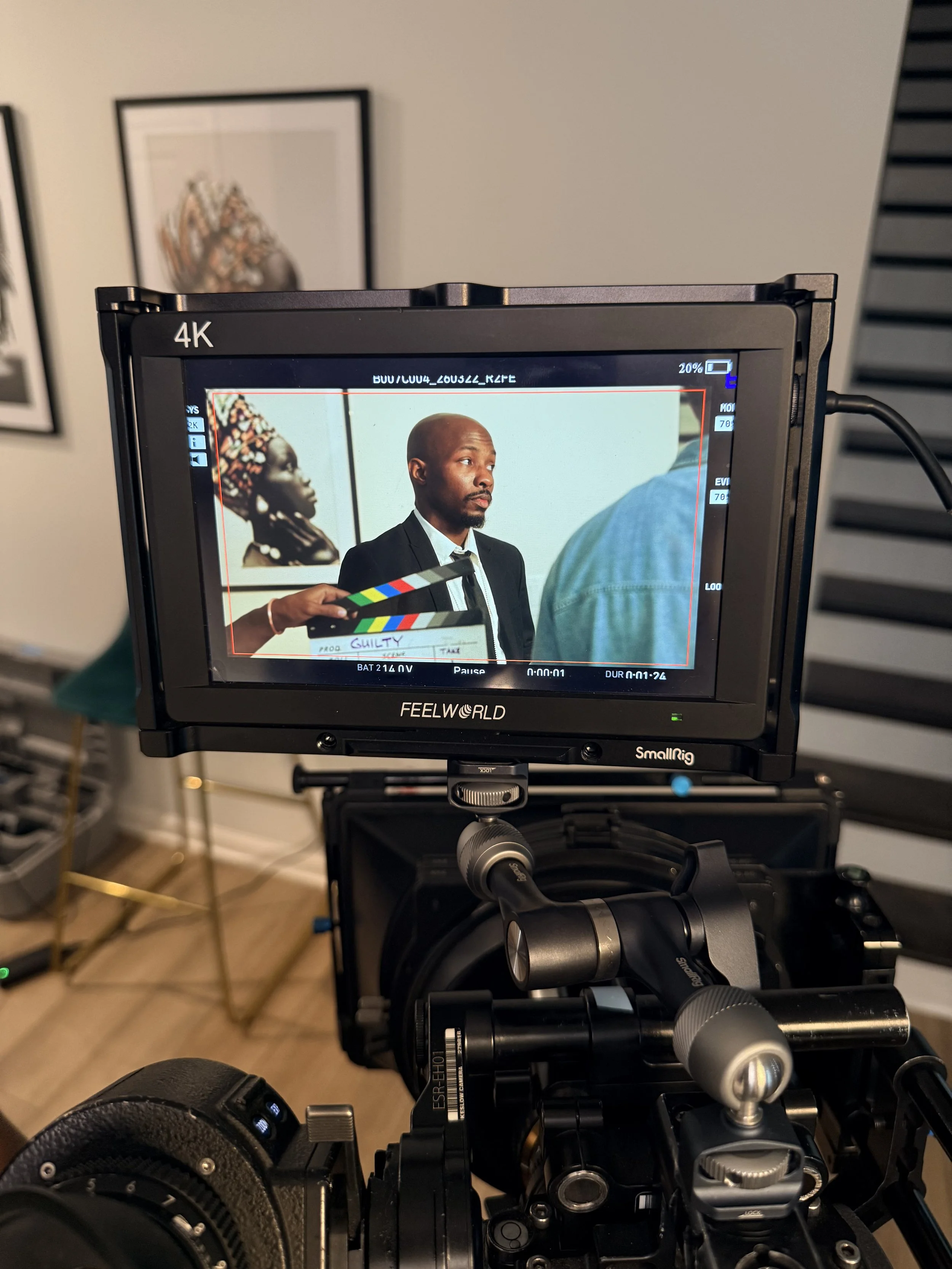 A camera on a tripod capturing a man with a shaved head and beard in a suit during a film shoot. In the background, a framed colorful Africa-inspired artwork is visible on a white wall.