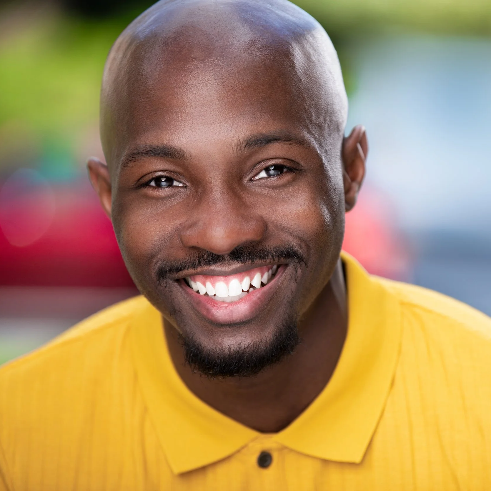 Close-up of a smiling man with a bald head, mustache, and beard, wearing a yellow shirt.