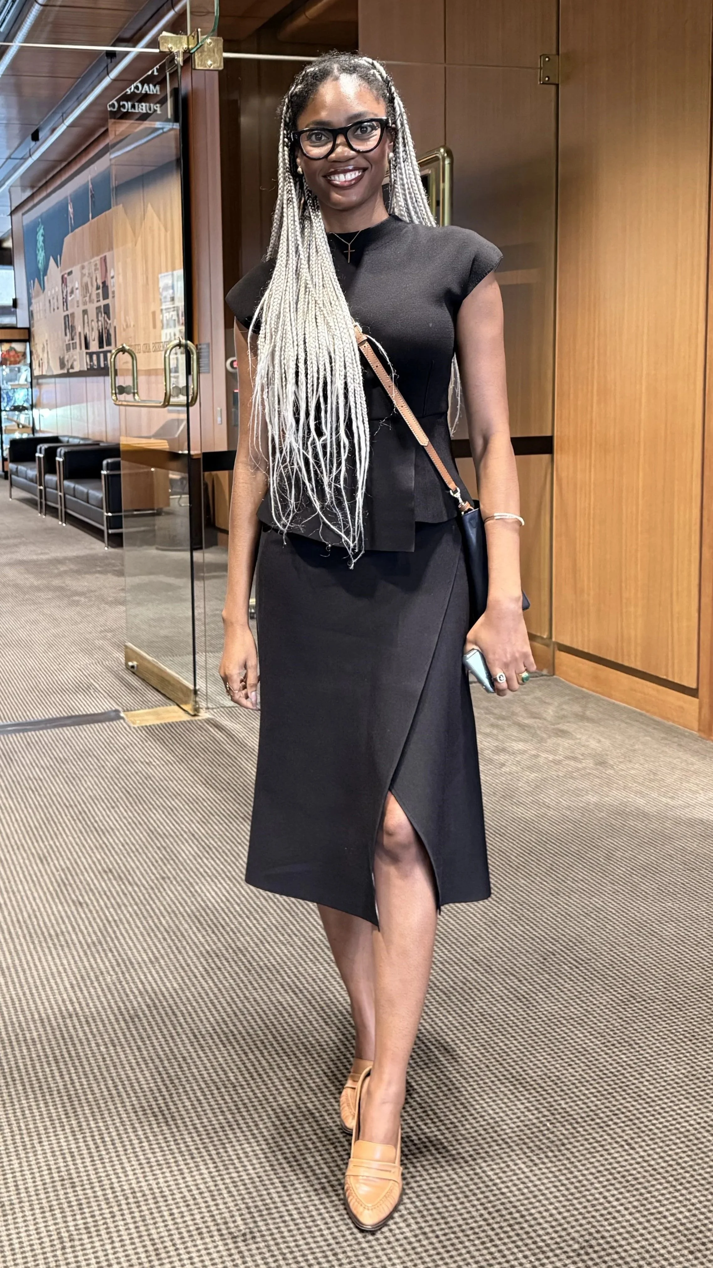 A woman with long braided hair, glasses, and dark skin, wearing a black top and skirt with a slit, tan shoes, and carrying a small bag, standing inside a building with wooden walls and glass doors.