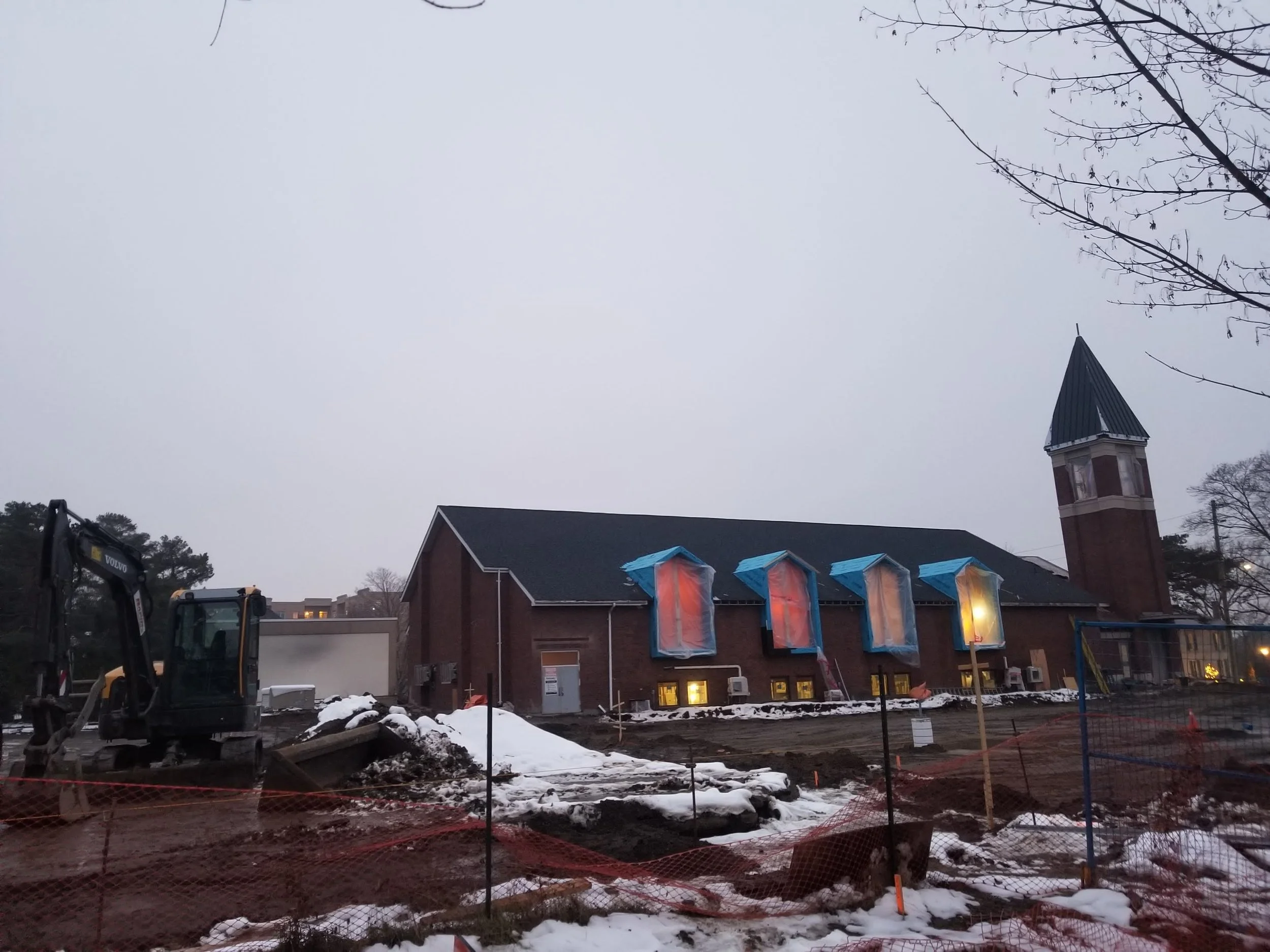 Construction site with dirt and snow, several small tents with colorful covers, a building with a steeple, and construction equipment including an excavator.