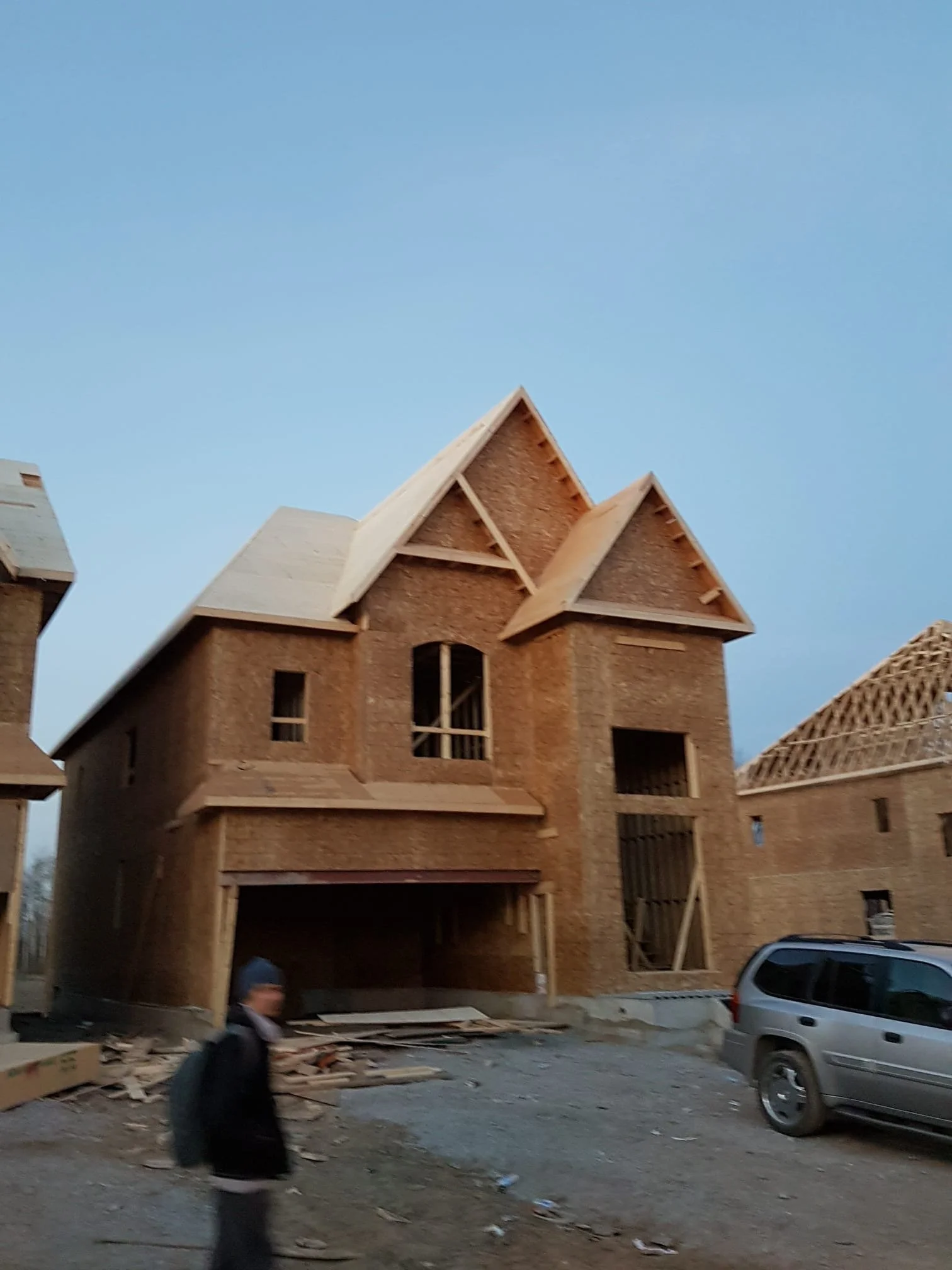 A house under construction with exposed wooden framing, located on a street with a parked vehicle in front and a person walking by. The sky is clear with no visible clouds.