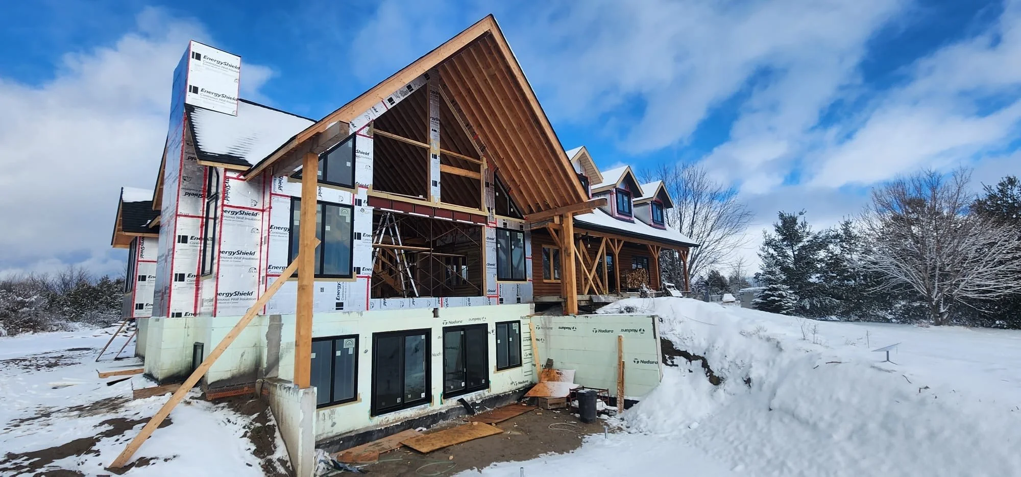 A house under construction in a snowy landscape with wooden framing, large windows, and a covered porch. The sky is blue with some clouds, and trees are in the background.