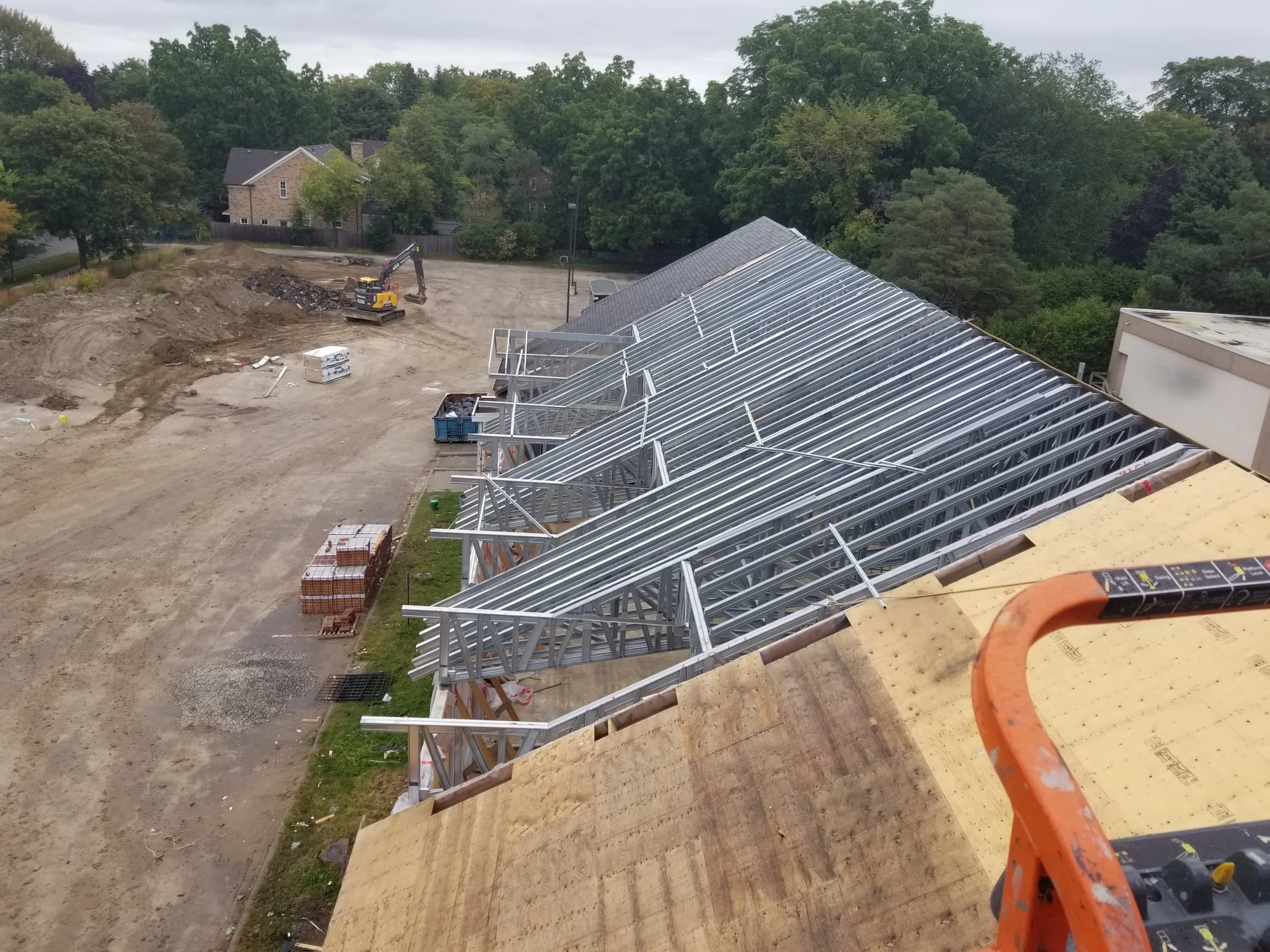 Construction site with metal framing for a building's roof, dirt piles, a small excavator, and wooden boards, surrounded by trees and residential houses in the background.