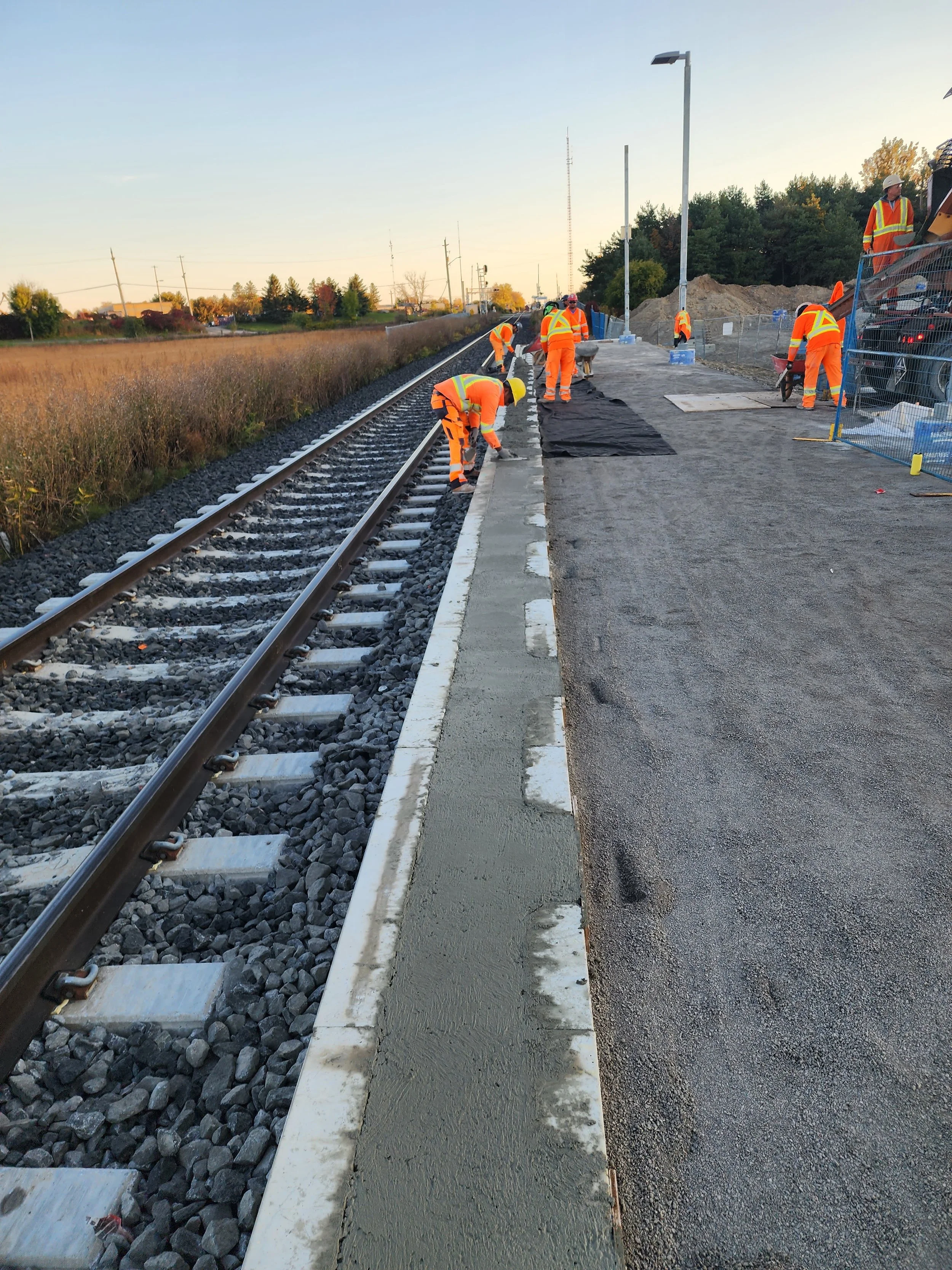 Road workers in orange safety vests and helmets working on a sidewalk beside train tracks, spreading concrete.