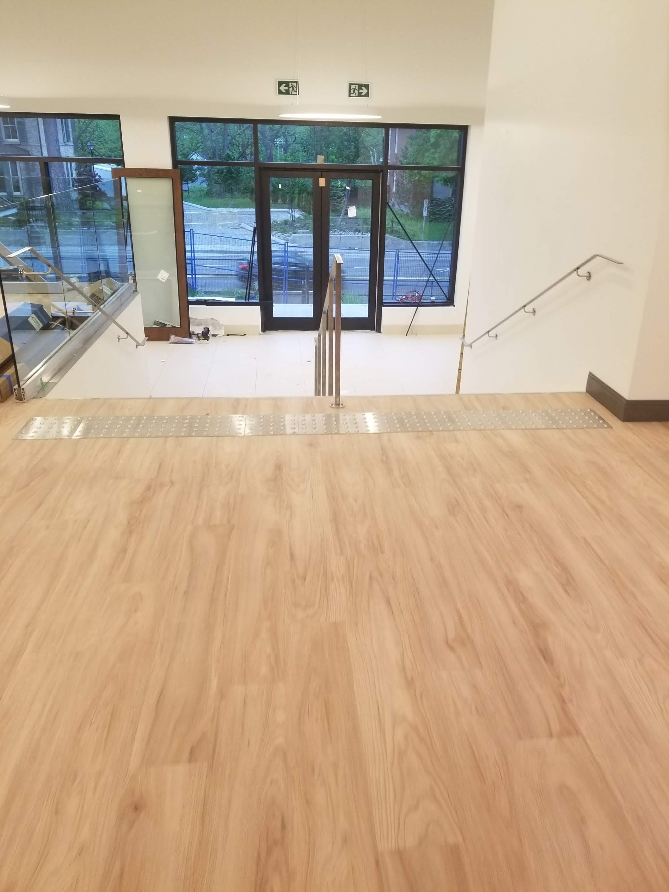 Interior view of a building staircase area with wooden floors, metal railings, glass display cases, and large windows showing outdoor greenery.