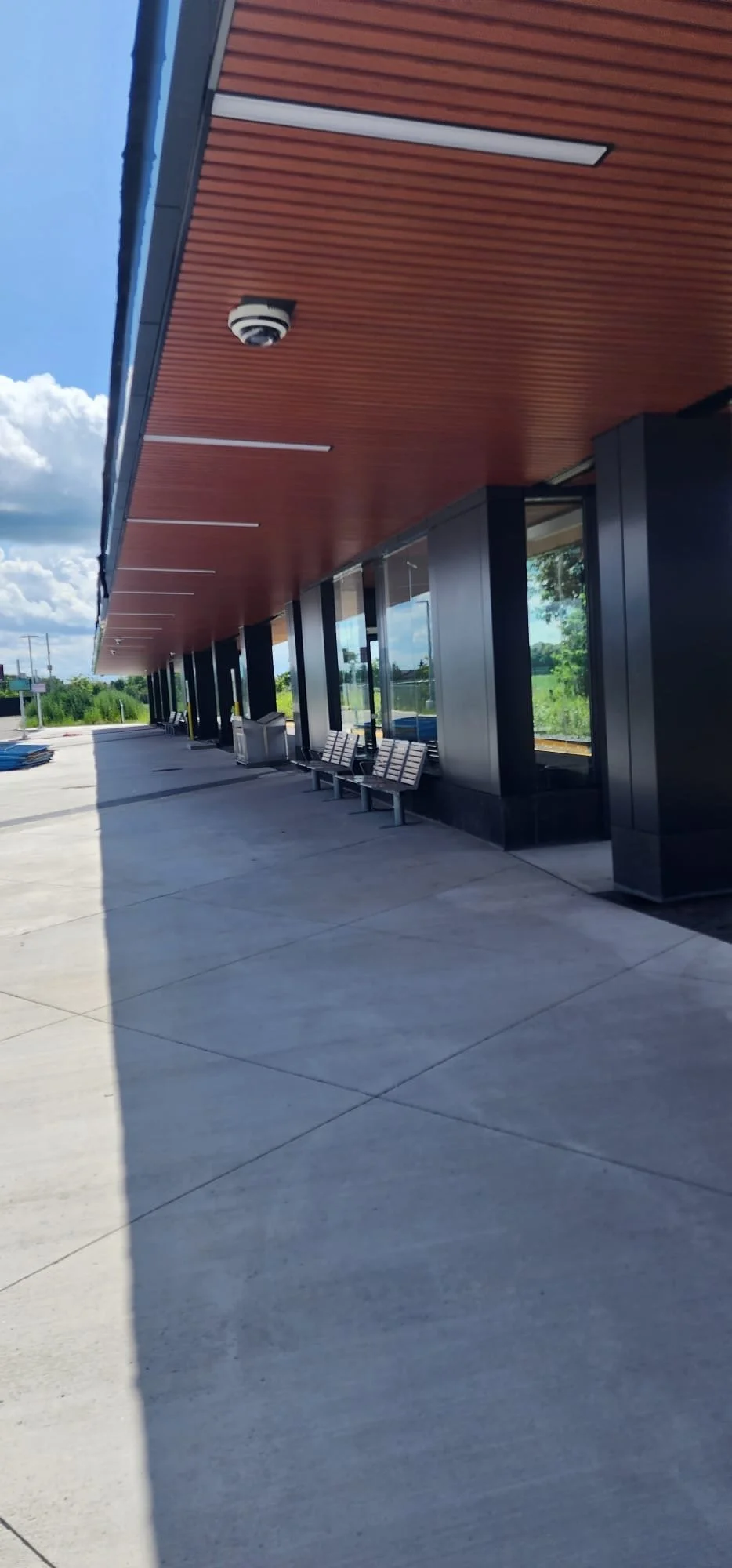 Empty sidewalk outside a modern building with large glass windows, benches, and a wooden ceiling overhang, under a blue sky with clouds.