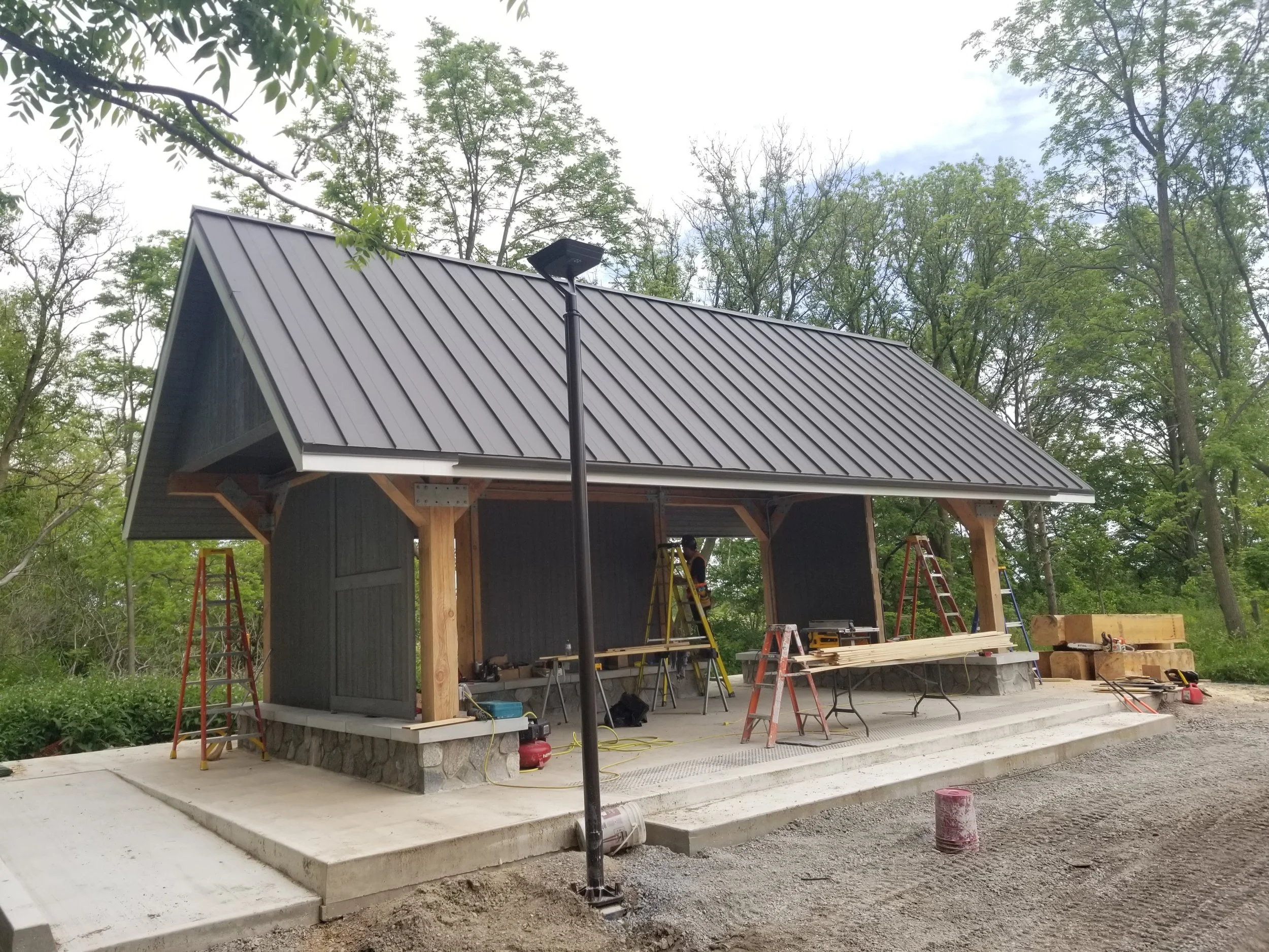 Construction site of a small building with a metal roof, surrounded by green trees, with ladders, tools, and building materials visible.