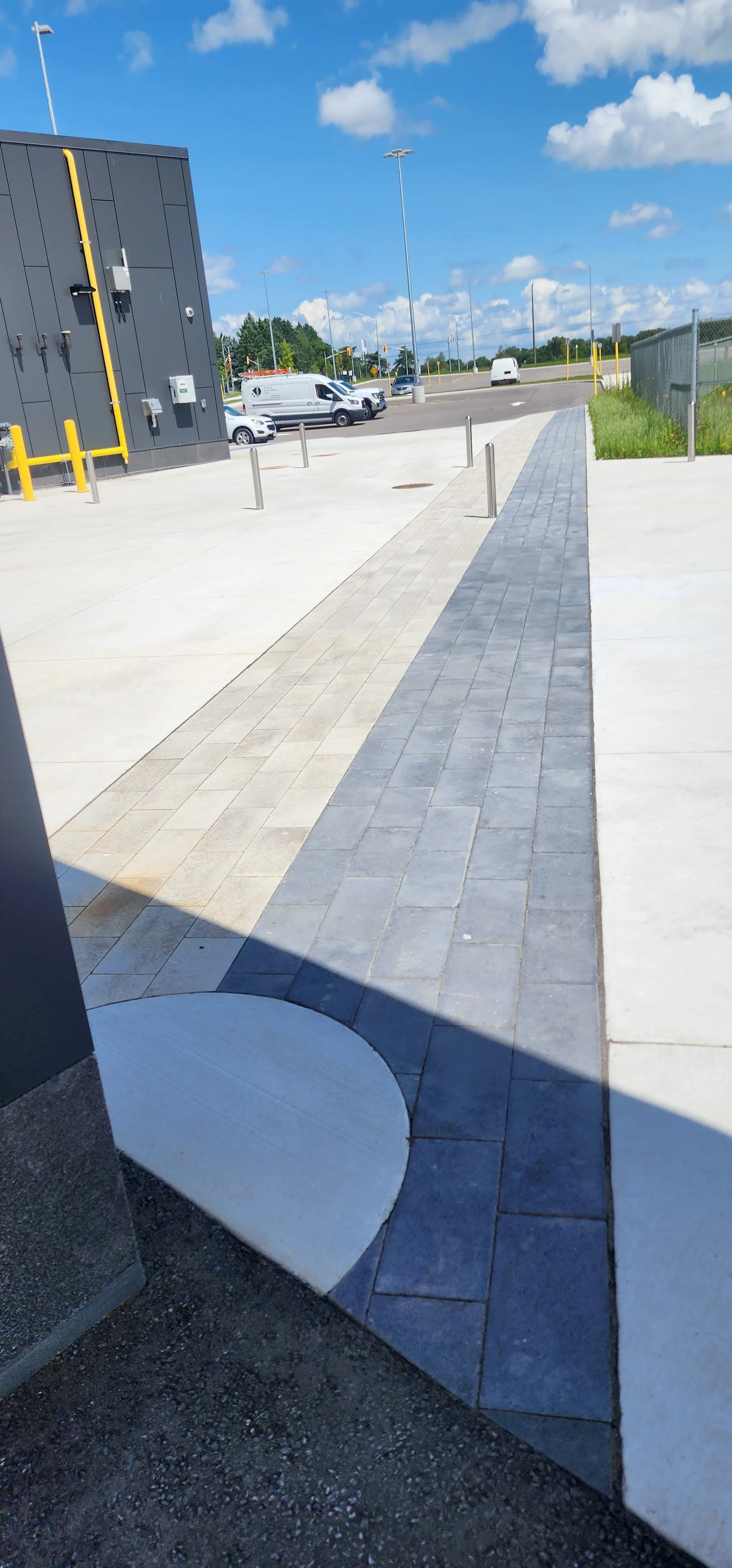 A paved sidewalk running alongside a black building with yellow pipes and utility boxes, with a parking lot and street with cars and vans in the background, under a partly cloudy blue sky.