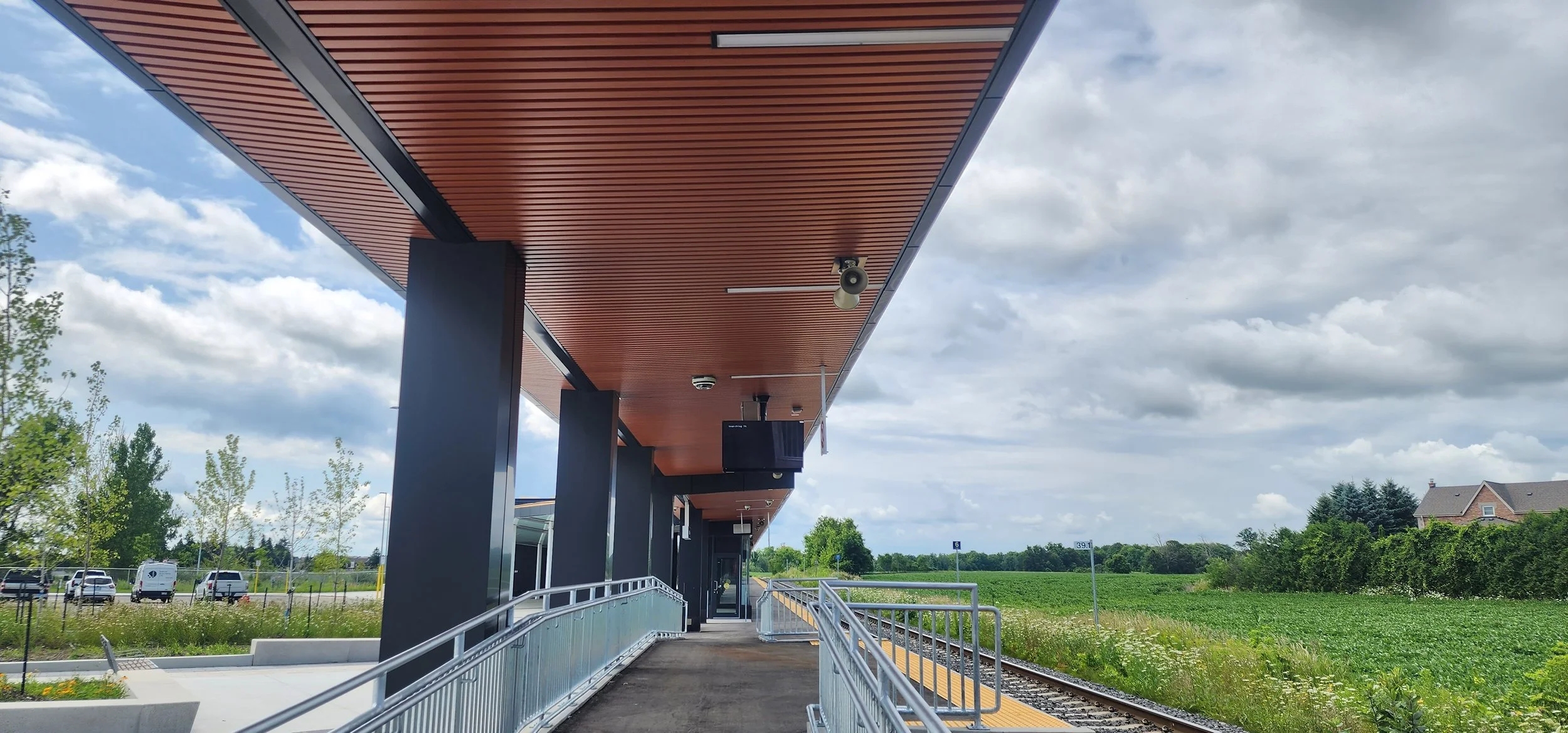 View of a modern train station platform with a covered roof, black supports, and a ramp with railings leading up to the station entrance. Railroad tracks run alongside the platform, with open green fields and a few houses in the distance. The sky is partly cloudy.