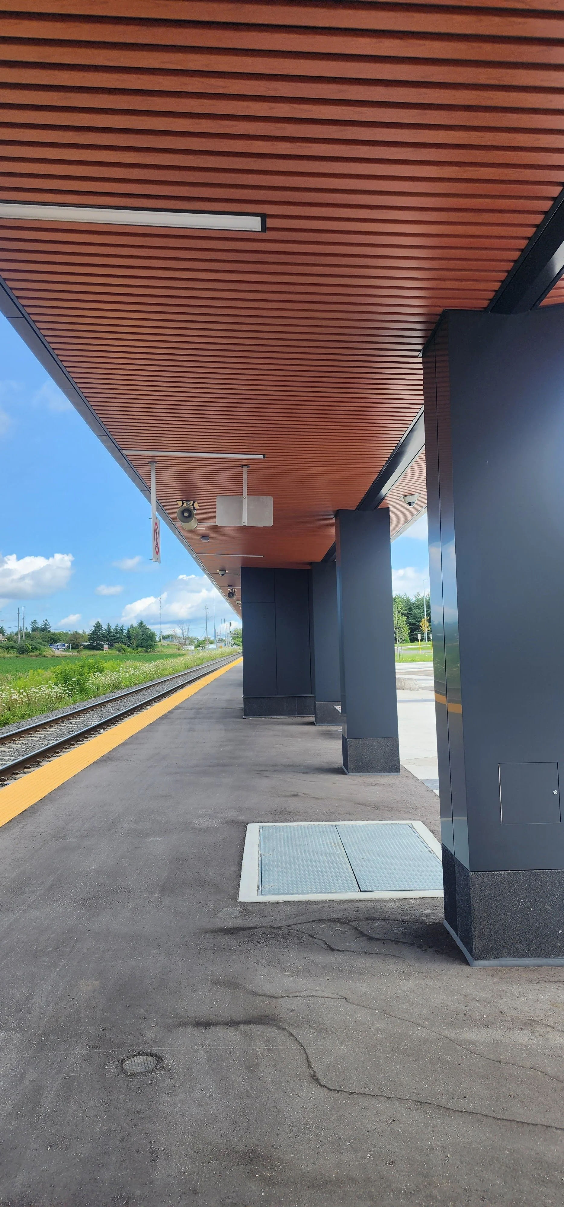 Empty train platform with a wooden roof, black columns, and a yellow safety line on the edge of the platform, next to train tracks under a partly cloudy sky.