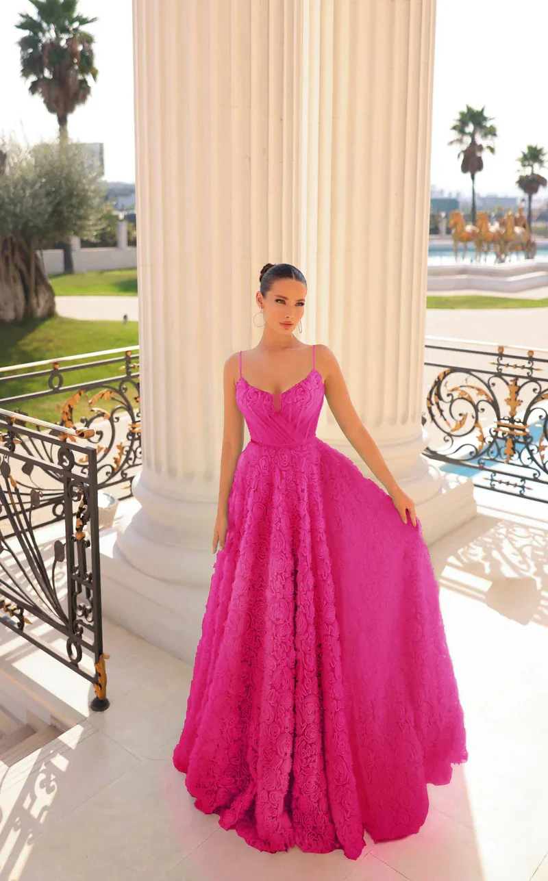 A woman in a bright pink, strapless gown with lace details, standing on a marble balcony with decorative iron railings and large columns, with palm trees and a fountain with golden horse sculptures in the background.
