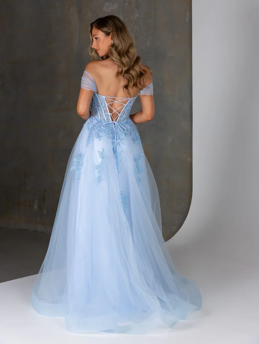 A woman with long wavy hair wearing a light blue off-the-shoulder wedding dress with lace-up back and sheer sleeves, standing against a textured gray background.