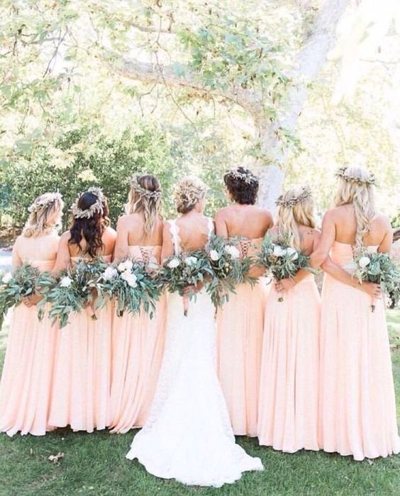A bridal party with seven women in peach dresses and wearing flower crowns, standing outdoors under trees, holding large green and white floral bouquets, with one woman in a white wedding gown in the center.