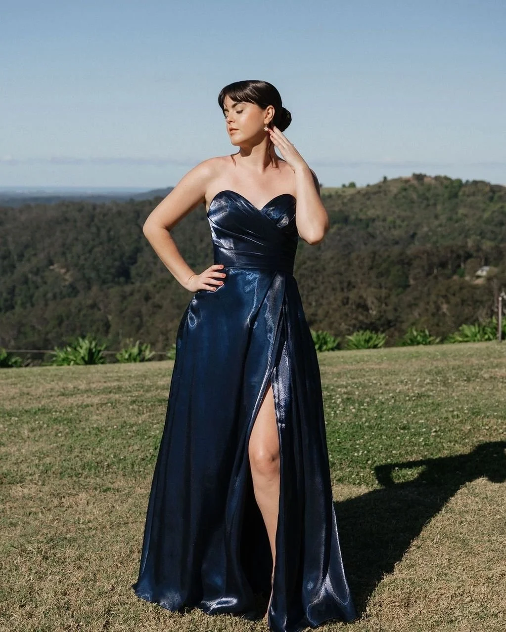 A woman in a strapless navy blue evening gown with a high slit standing outdoors on grass with a scenic background of hills and trees.