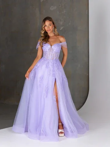 A woman in a lavender off-the-shoulder ball gown with a high slit, standing against a mixed background of grey and white walls.