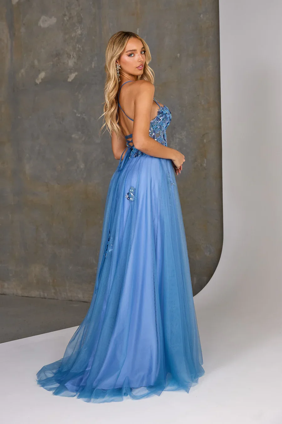 A woman with long blonde hair in a blue evening gown with floral embroidery, standing with her back slightly turned and looking over her shoulder against a neutral backdrop.