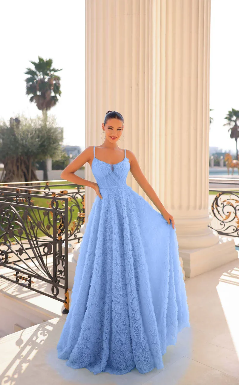 A woman in a light blue, embroidered, floor-length gown with spaghetti straps standing on a balcony with ornate black and gold railing. She is smiling and posing with one hand on her hip. Palm trees are visible in the background.