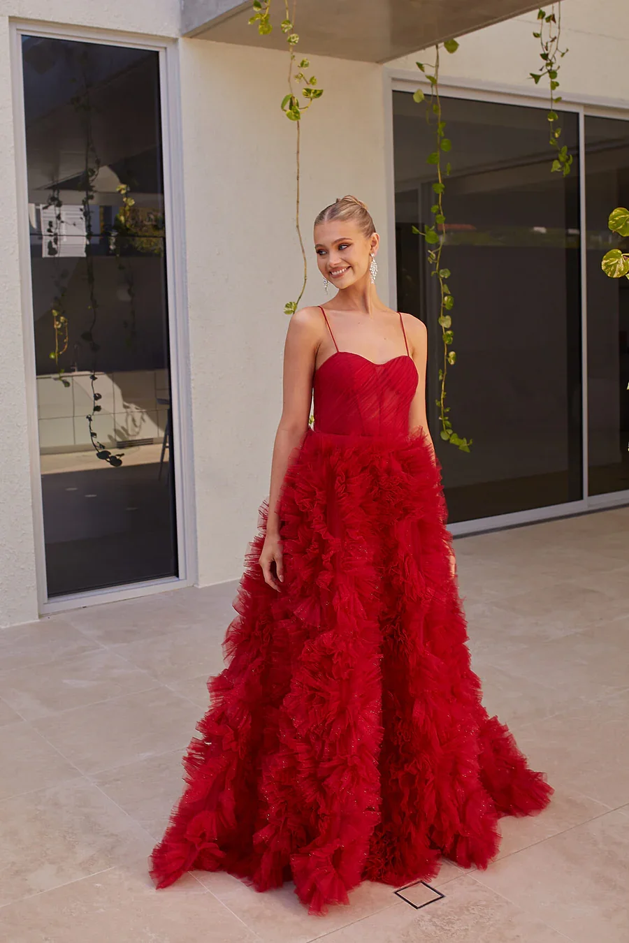 A young woman in a red, ruffled evening gown standing on a tiled patio near sliding glass doors and hanging plants, smiling.