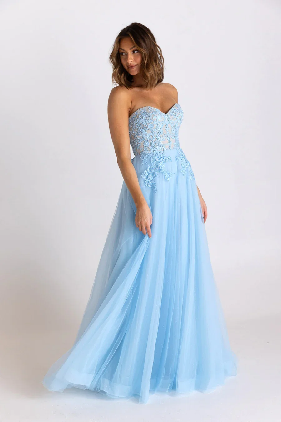 A woman wearing a light blue strapless dress with lace detailing on the bodice, standing against a plain white background.