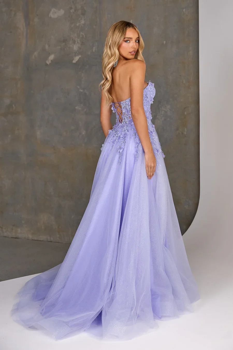 A woman in a lavender strapless wedding dress with lace and embroidery details, posing in a studio with a gray background.