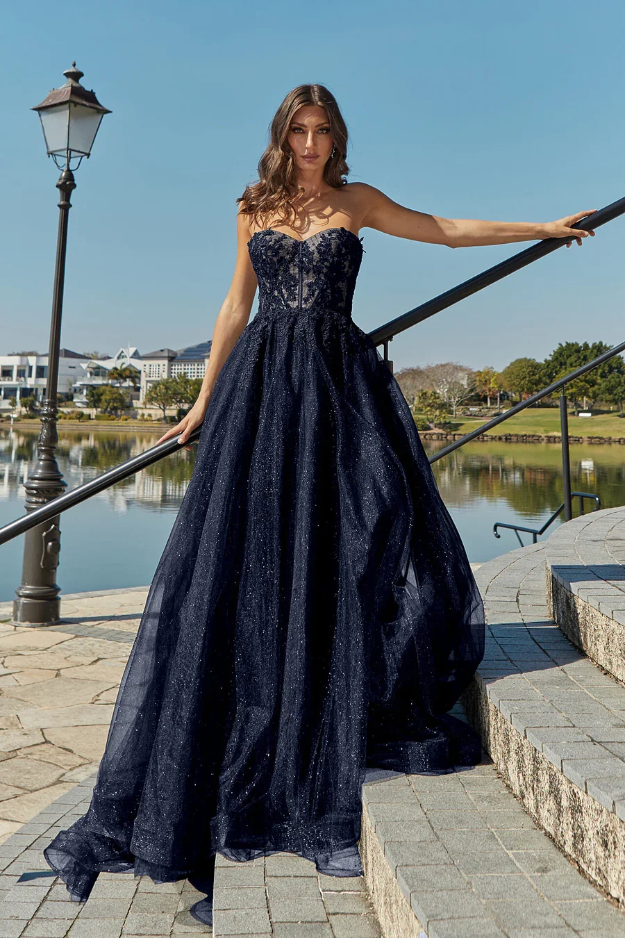 A woman in an elegant, strapless, dark blue gown with glittery fabric, posing on a stone staircase by a lake.