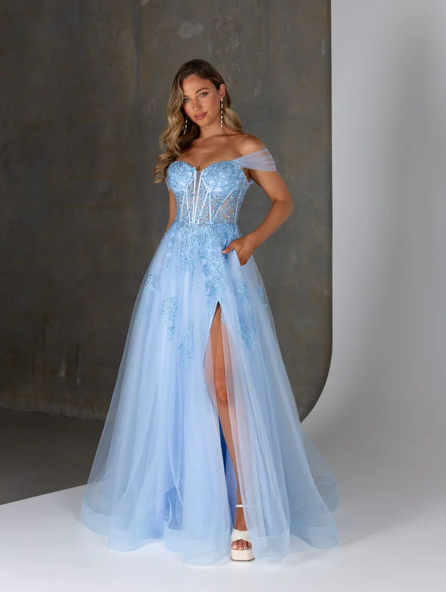 Young woman in a light blue lace off-the-shoulder gown with a slit, standing in front of a textured wall and a plain white background.