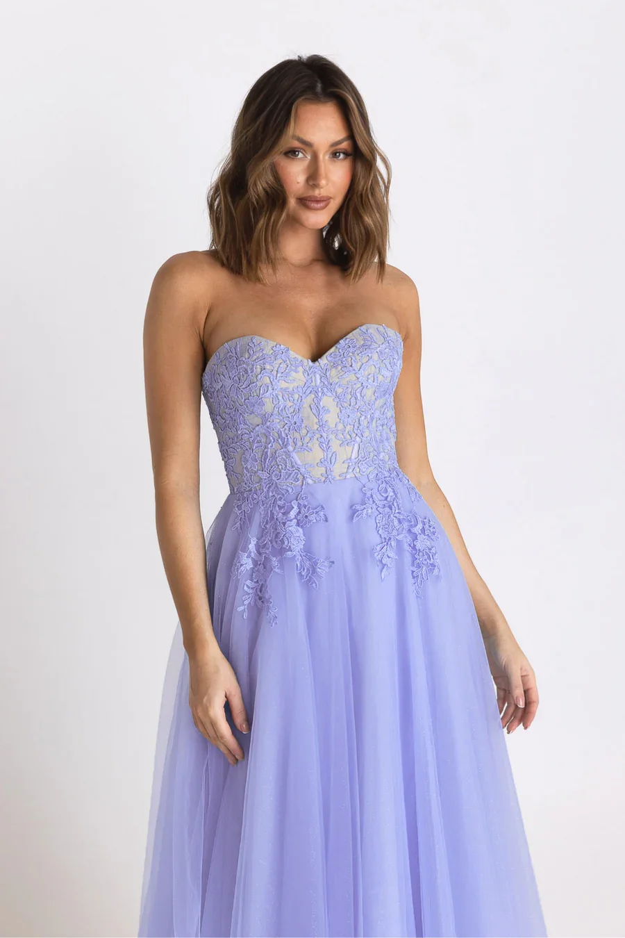 A woman in a strapless lavender lace and tulle gown standing against a plain white background.