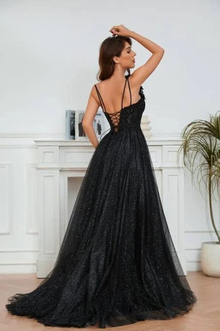 A woman in a black, sparkly evening gown with spaghetti straps, standing in a room with white paneling and a fireplace, with a potted plant on the right.