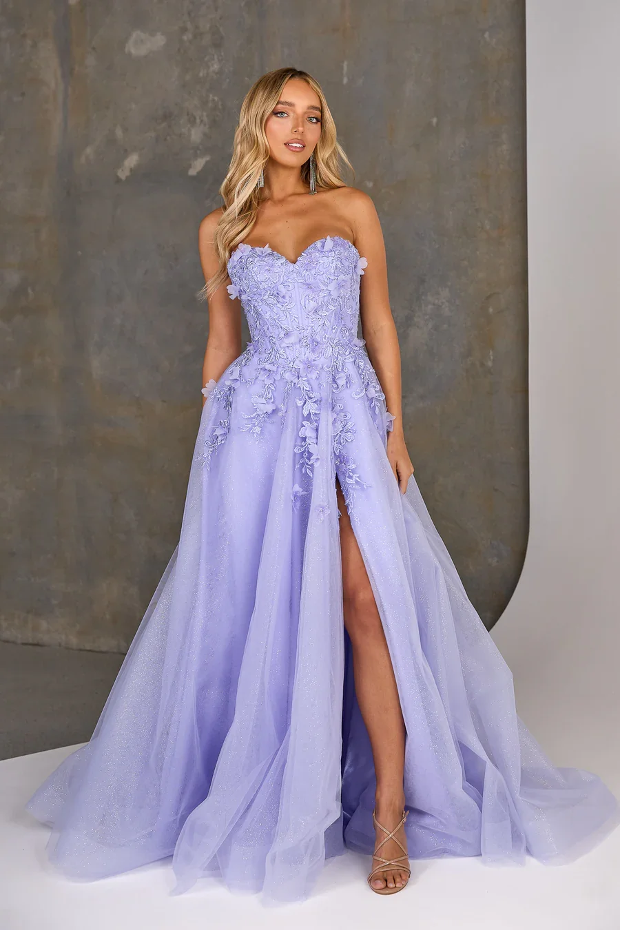 A woman wearing a strapless lavender wedding gown with floral embroidery and a high slit, standing in front of a gray wall.