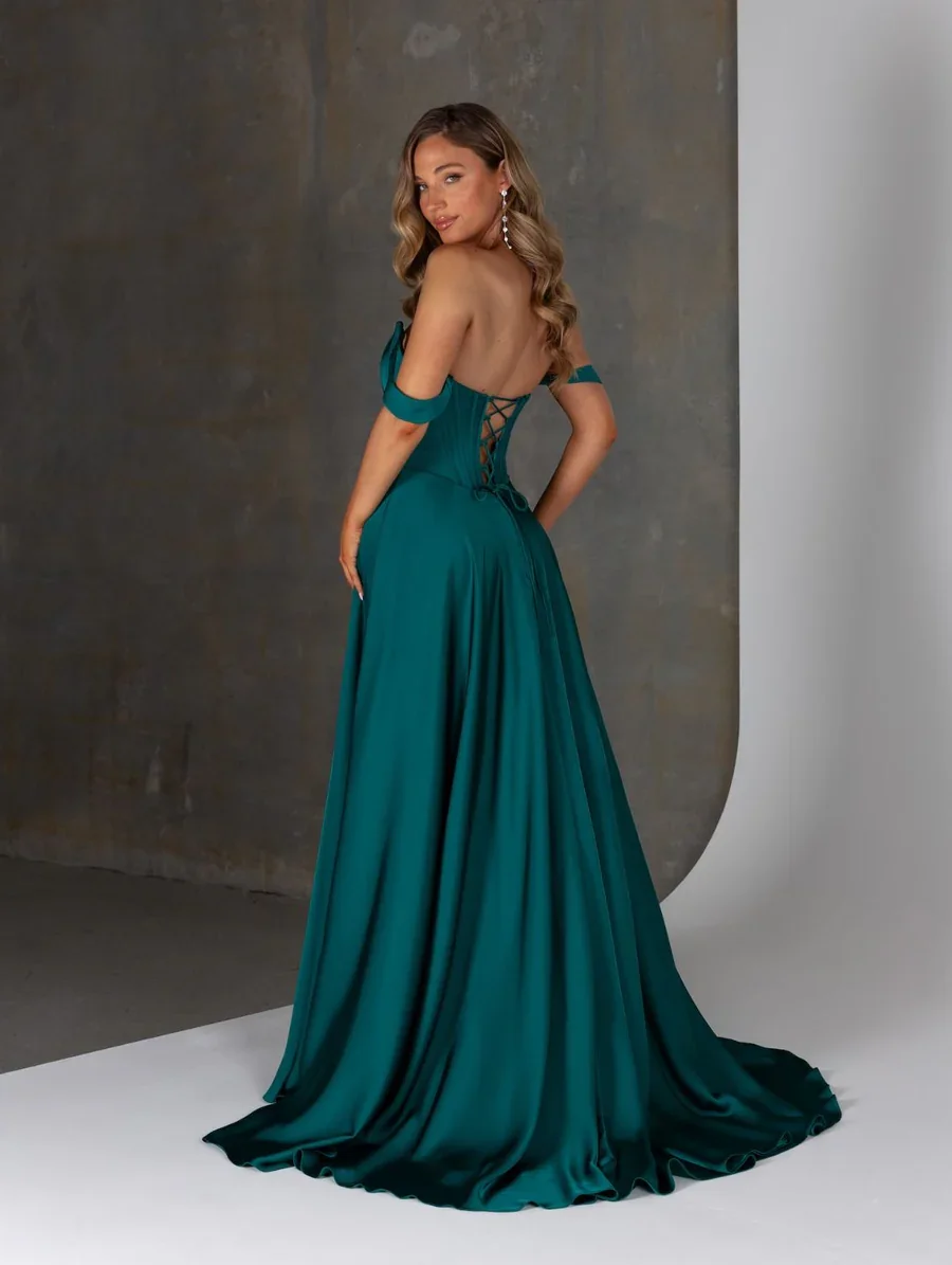 A woman in a teal satin gown with a lace-up back, off-shoulder sleeves, and long flowing skirt, standing against a textured gray and white background.