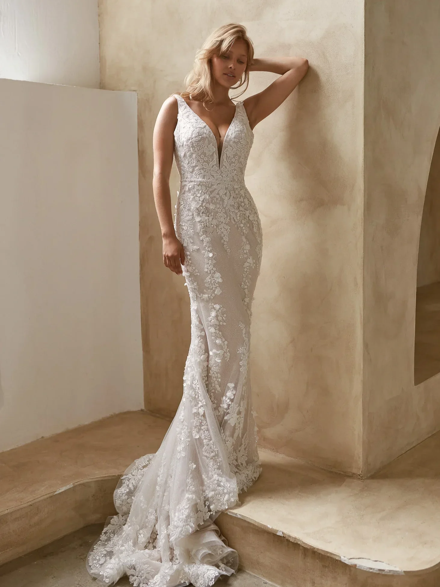 A woman in a white lace wedding dress standing on a stone staircase, with one arm raised behind her head and a neutral expression.