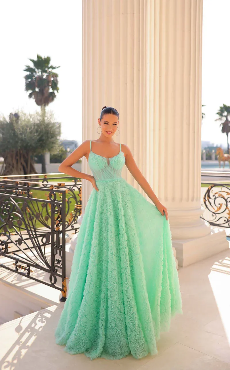 A young woman wearing a light green lace dress with spaghetti straps, standing on a terrace with marble columns and ornate black and gold railings, palm trees in the background, smiling at the camera.