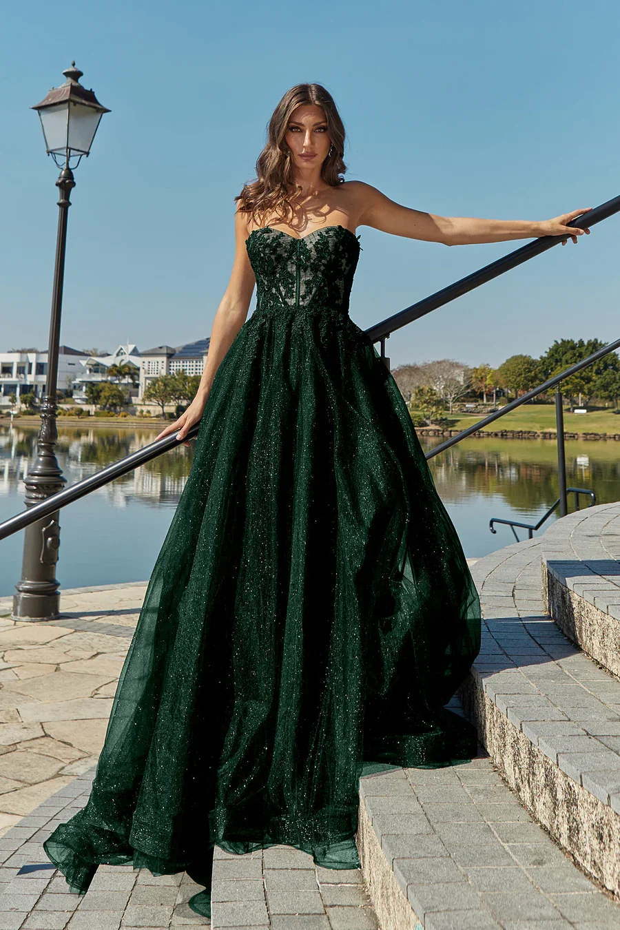 A woman in an elegant, strapless, dark green gown with a glittery tulle skirt, standing outdoors on stone steps by a lake with buildings and trees in the background.
