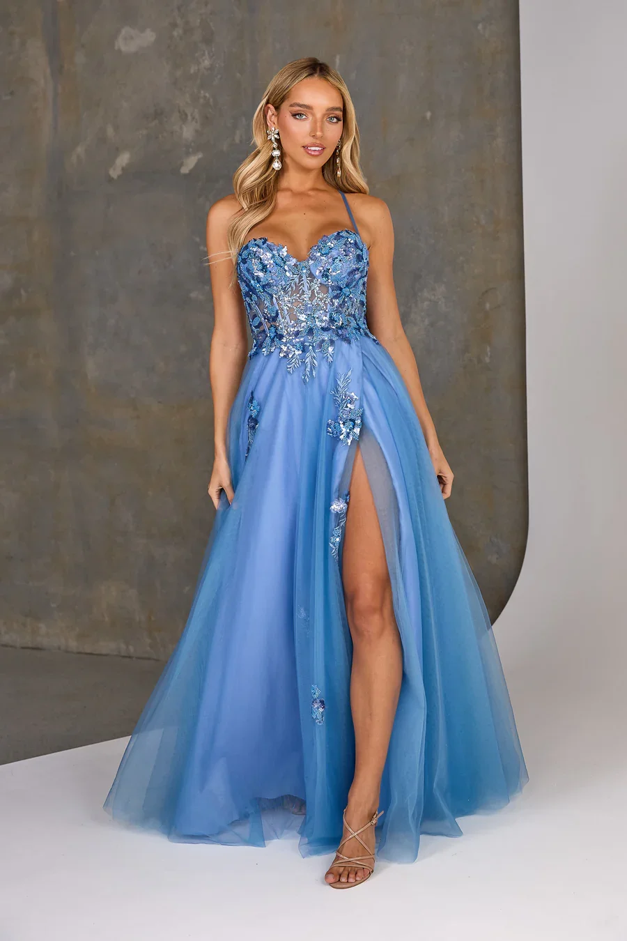 A young woman wearing a blue evening gown with floral embellishments, a thigh-high slit, and spaghetti straps, posing in front of a grayish textured wall, with long blonde hair, earrings, and strappy high heels.