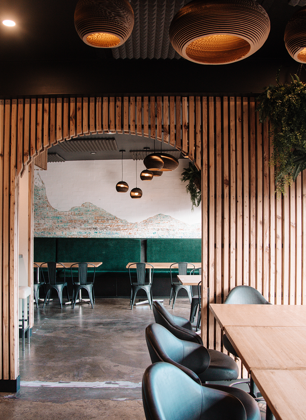 Interior of a modern cafe or restaurant with wooden slat dividers, black and green seating, hanging pendant lights, and a brick wall with a mountain mural.