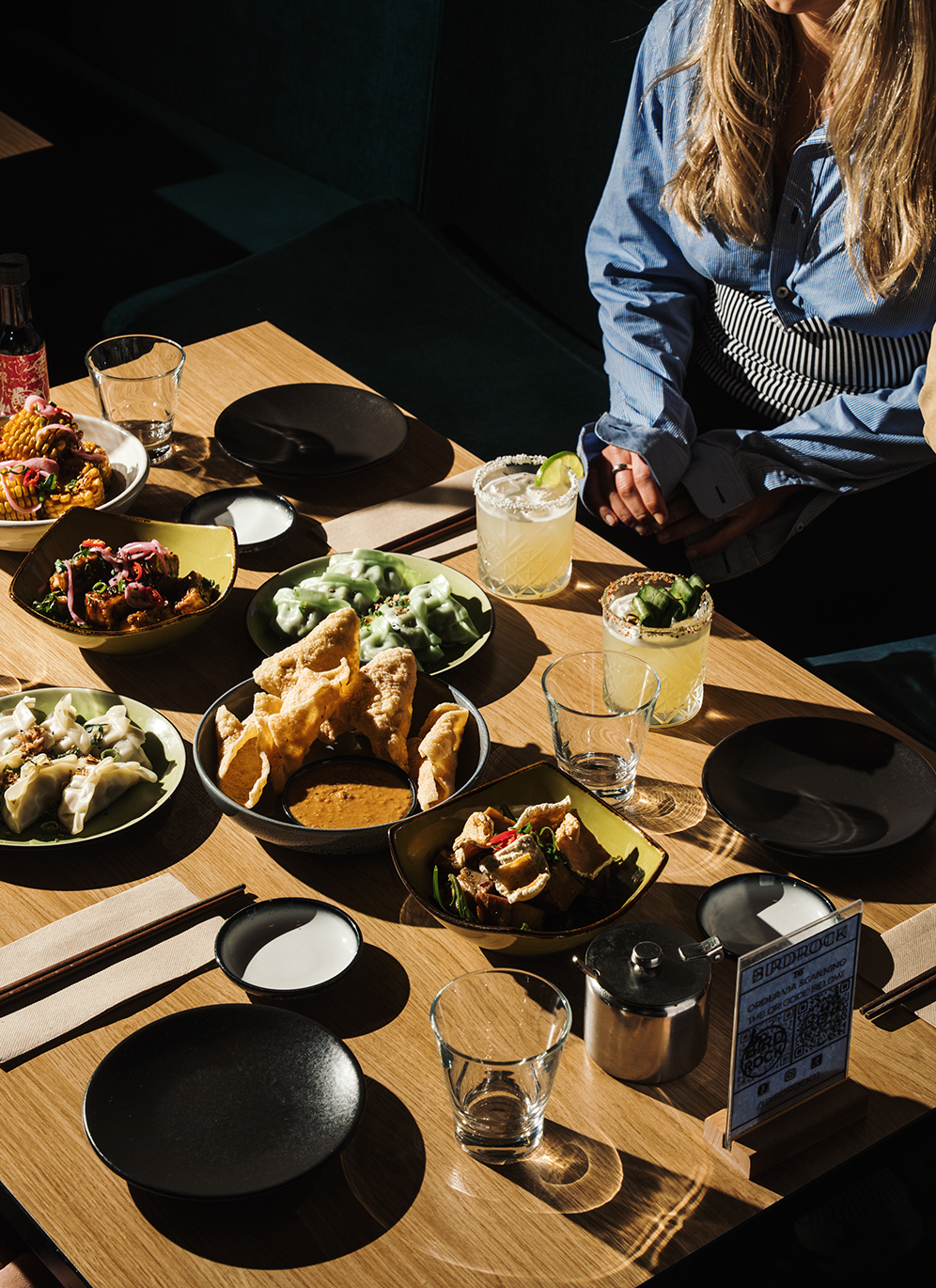 A wooden dining table set with various dishes including dumplings, salads, fried chicken, and beverages, with a person sitting nearby.