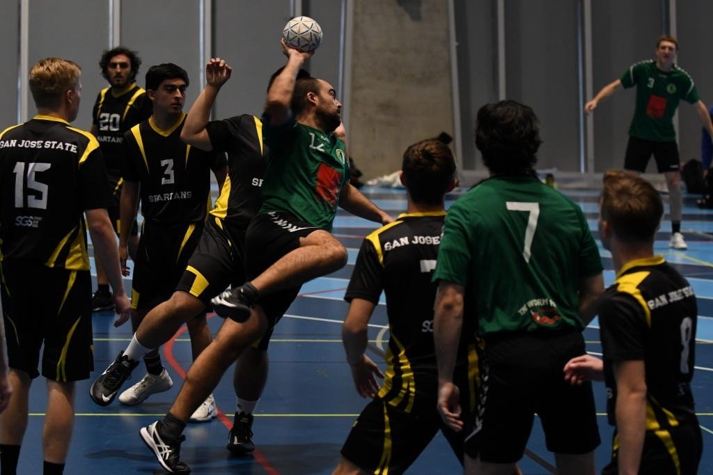 Indoor men's volleyball game with players in black and yellow uniforms and green uniforms; one player in green jumping to hit the ball near the net, while others watch and prepare to react.